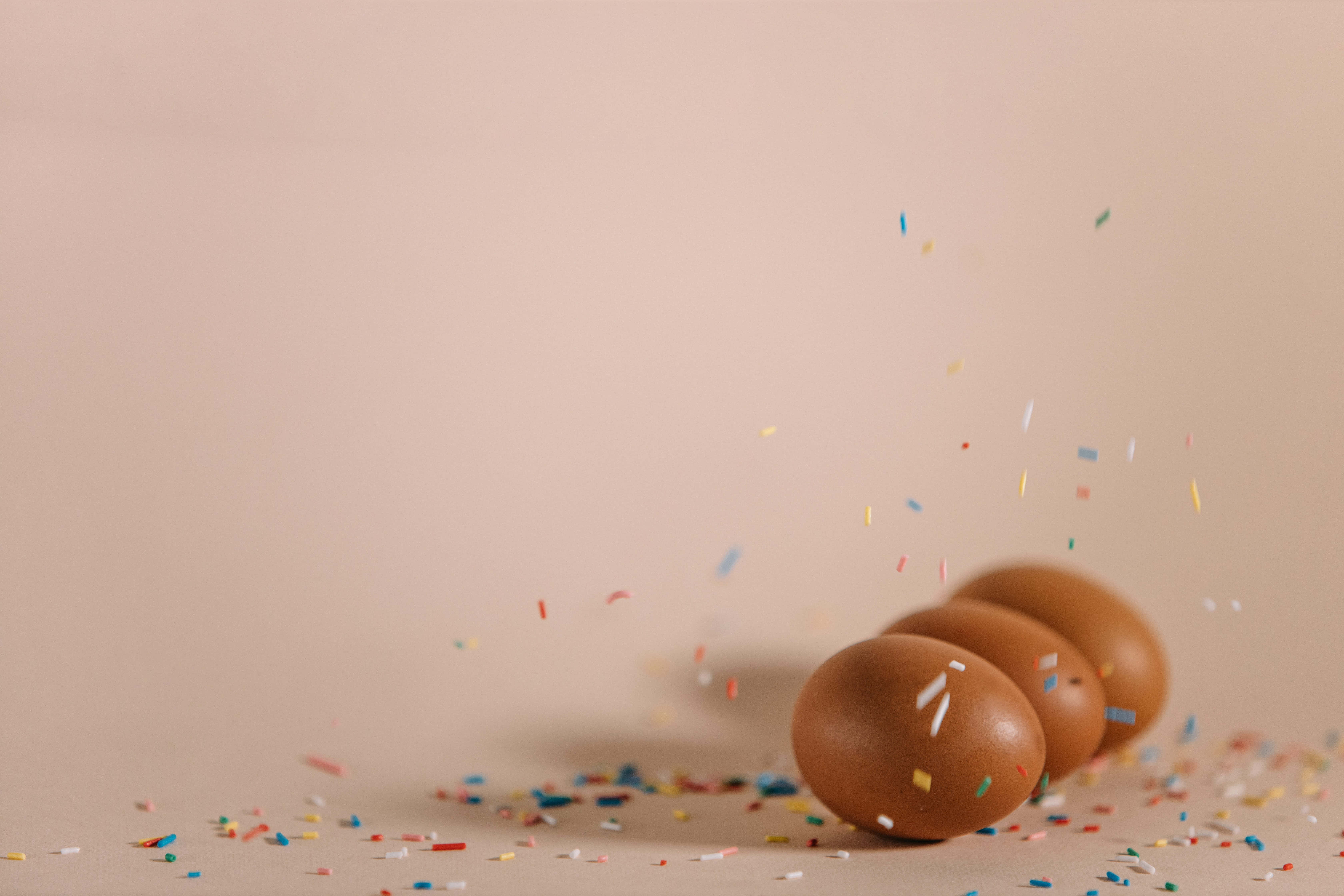 Brown Eggs 桌布 壁紙 — 四隻棕色雞蛋在米色表面上滾動，彩色紙屑以慢動作在它們周圍爆開。