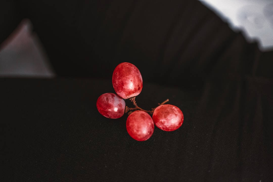 四顆紅葡萄 壁紙 — 四顆飽滿的紅葡萄在柔和的燈光下，在深色紋理表面上閃閃發光。