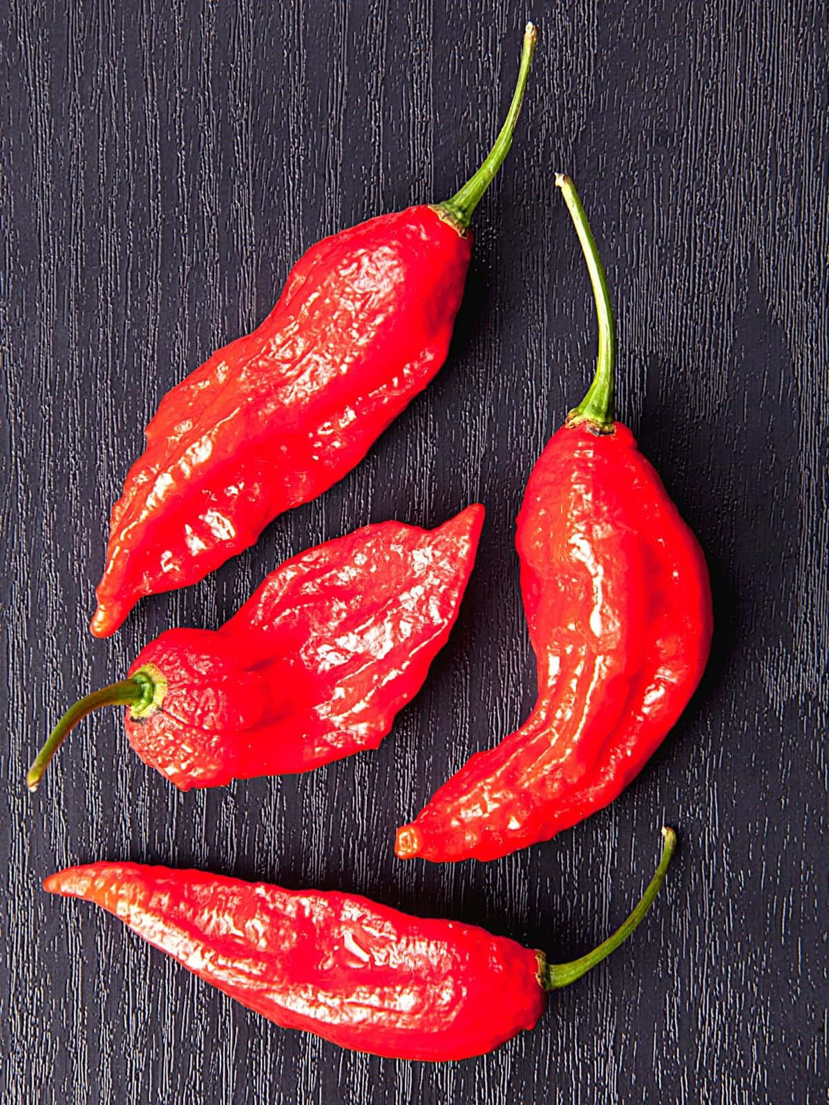 Ghost Pepper 桌布 壁紙 — 四顆鮮紅的哈瓦那辣椒，帶有綠色莖部，放置在深色紋理木質表面上。