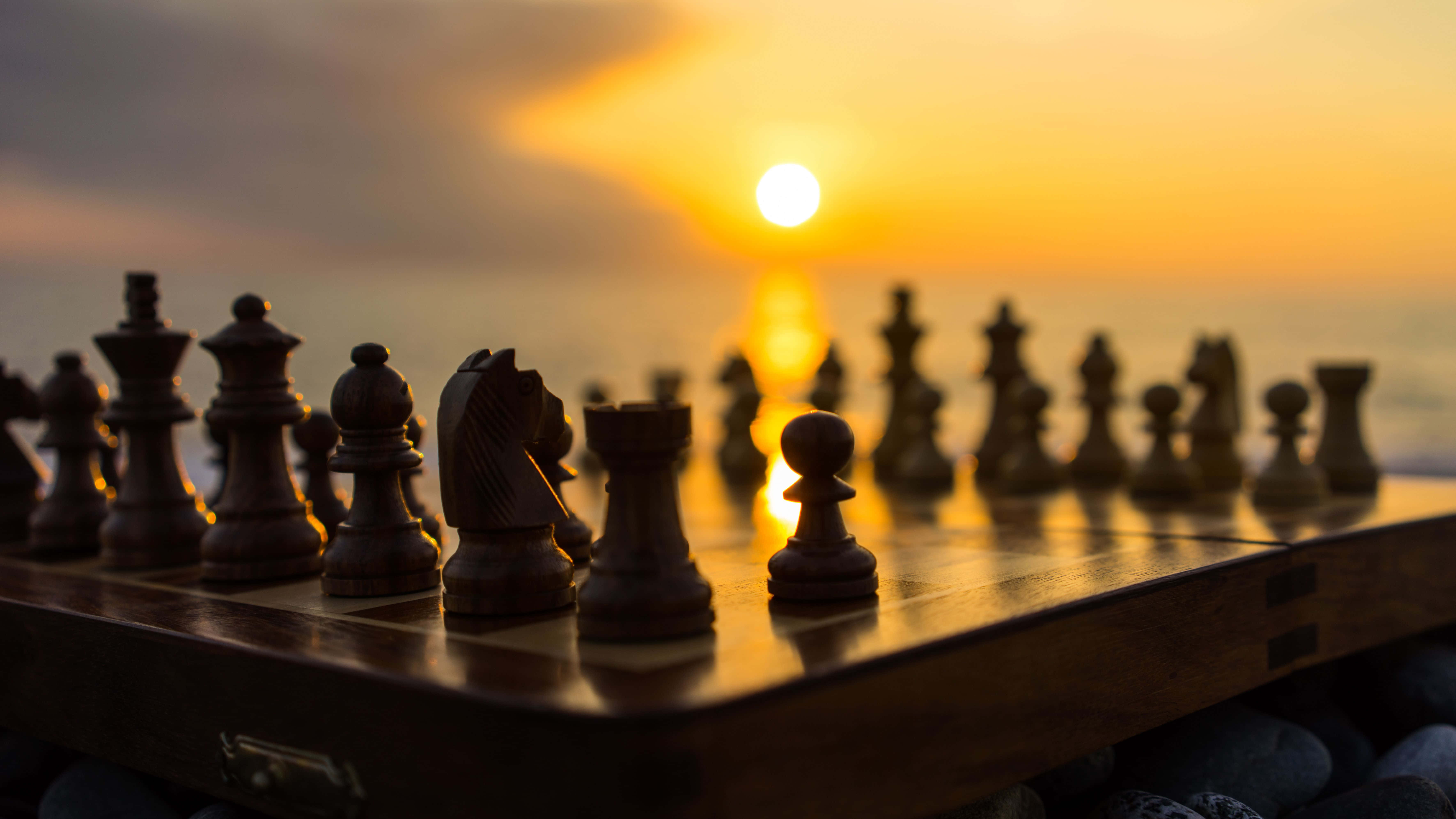 國際象棋 桌布 壁紙 — 國際象棋棋子以剪影形式立於木製棋盤上，夕陽在它們後方的大海上落下。