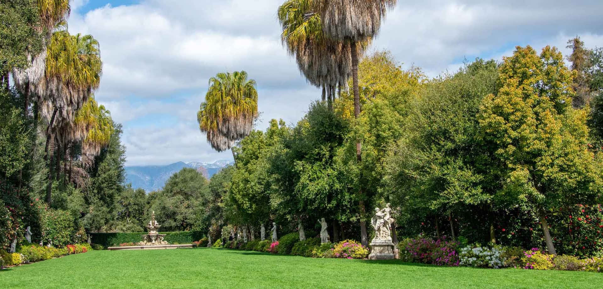 園丁打造美麗戶外花園景觀 壁紙 — 一個宏偉的花園，擁有茂密的綠色草坪、棕櫚樹和古典雕像，天空部分多雲，遠山可見。