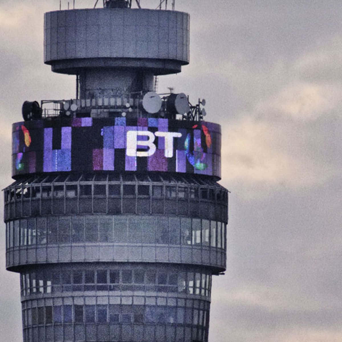 圓形螢幕包圍的BT塔手機壁紙 壁紙 — 一座高聳的BT建築在陰沉的灰色天空中展示了一個充滿活力的數碼彩虹標誌。
