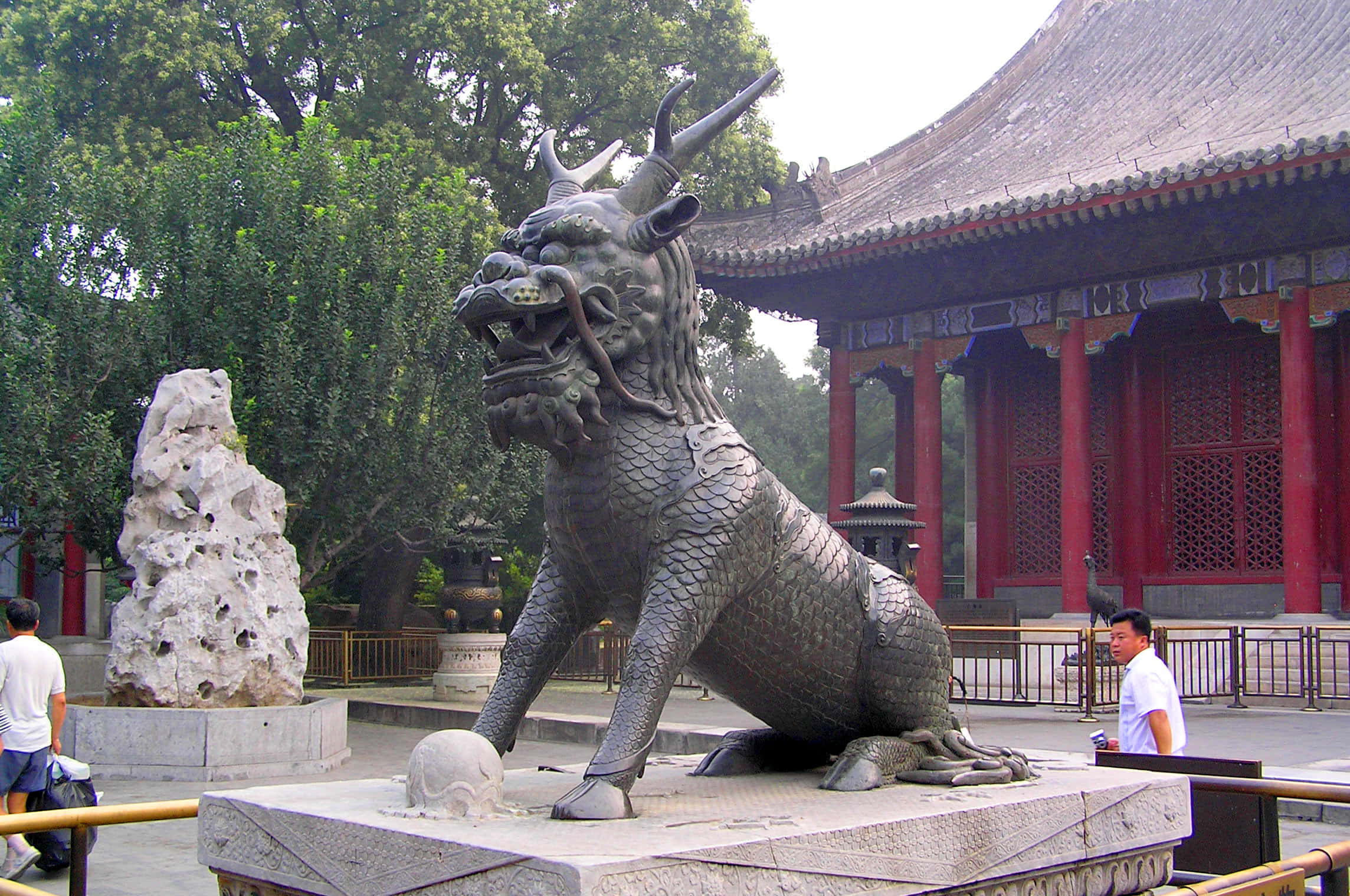 圓明園內的麒麟雕像 壁紙 — 一個大型金屬麒麟雕像守護著傳統中國寺廟庭院，庭院內有紅色柱子和茂密的綠色樹木。