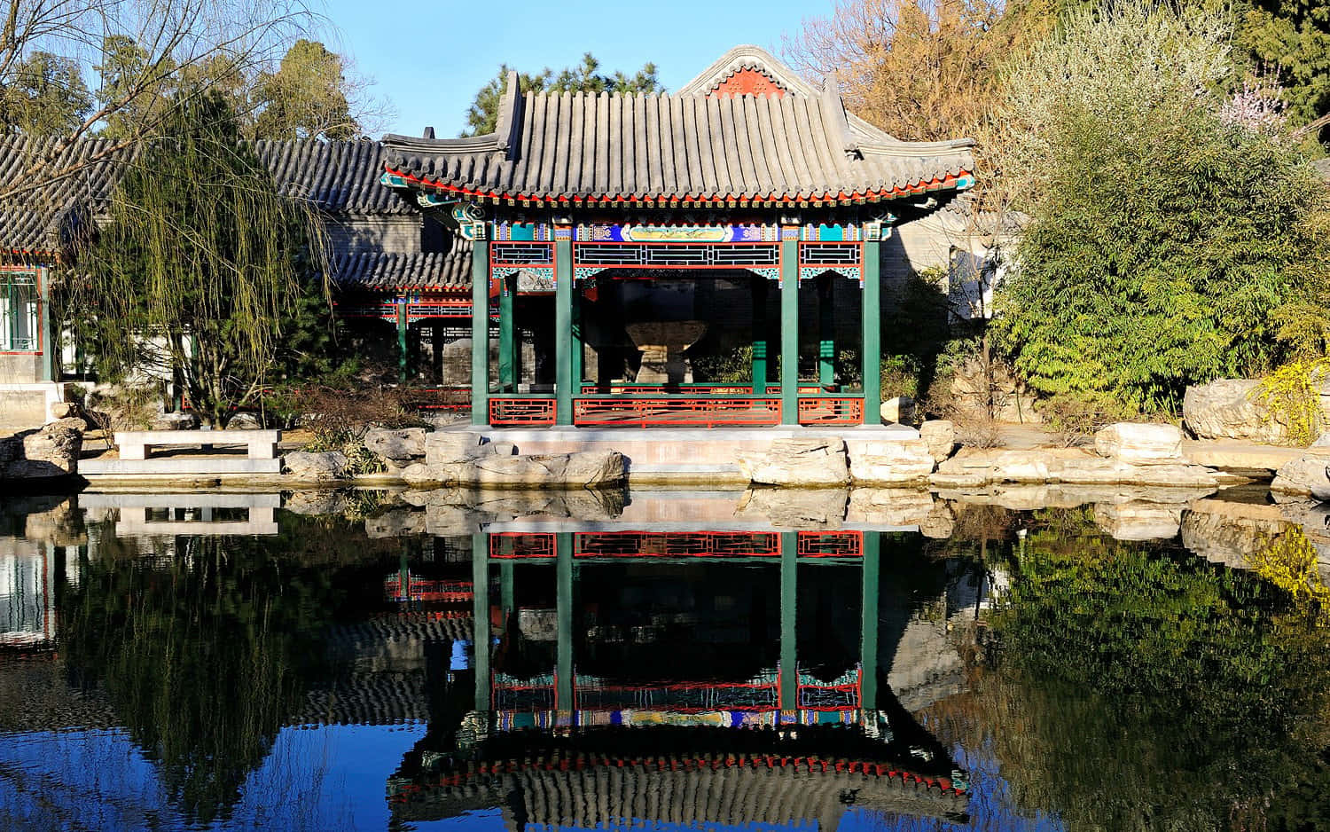 圓明園園林池塘 壁紙 — 一座傳統中式亭閣，綠色柱子配紅色點綴，佇立於平靜池塘旁，在清澈藍天下倒映出其精緻細節。