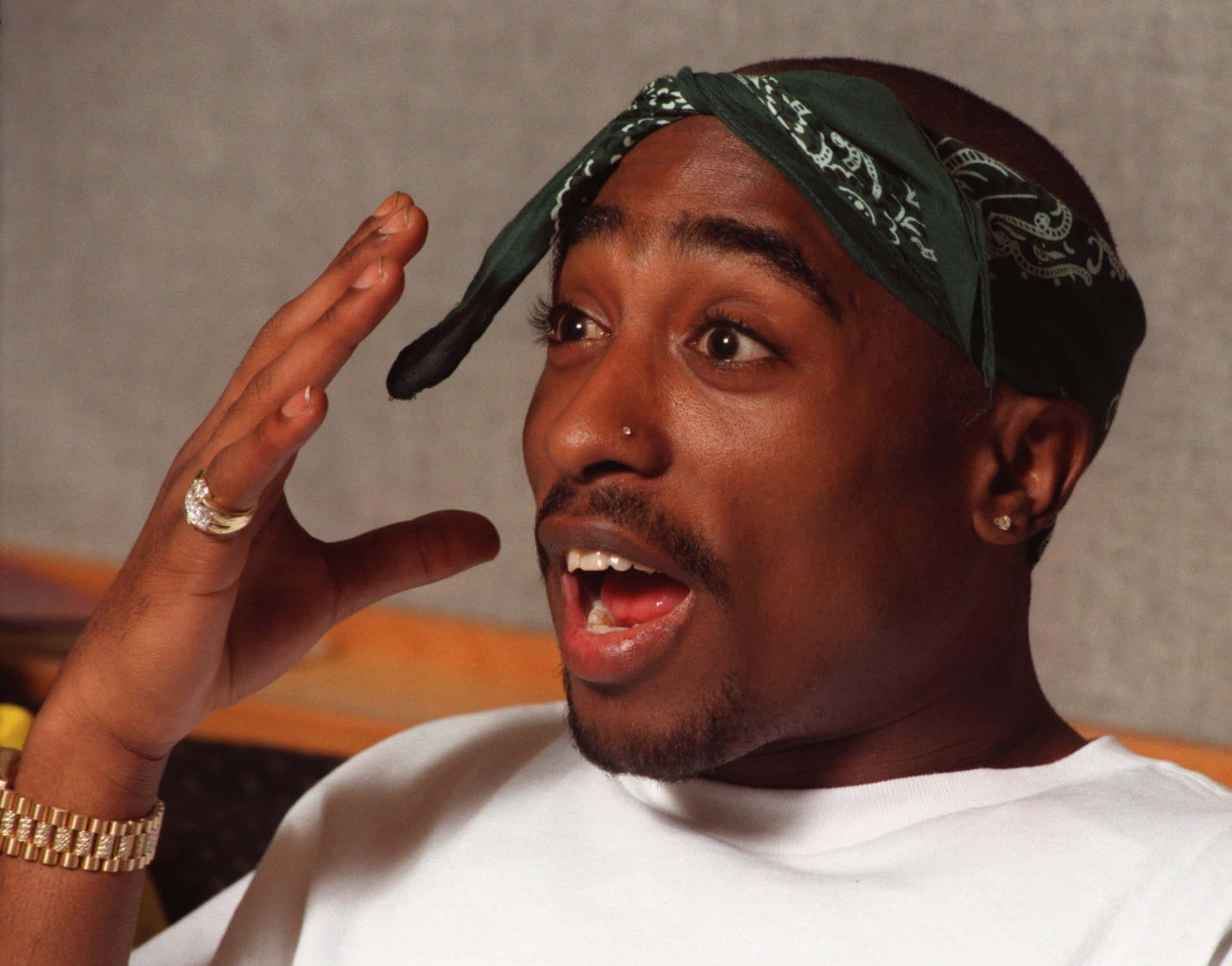 Tupac Shakur 身穿白襯衫，戴著綠色頭巾和金飾，做出驚訝的表情。