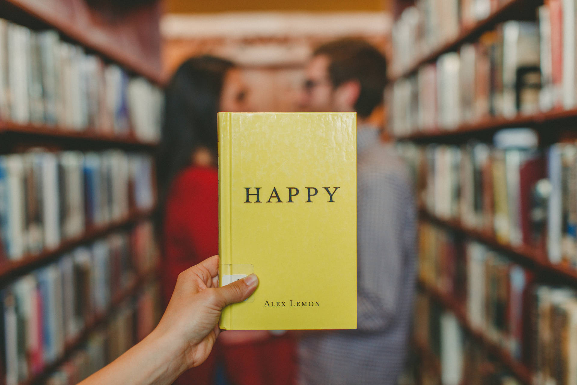 手拿一本名為「HAPPY」的黃色書在圖書館中，後面有一對情侶在接吻。