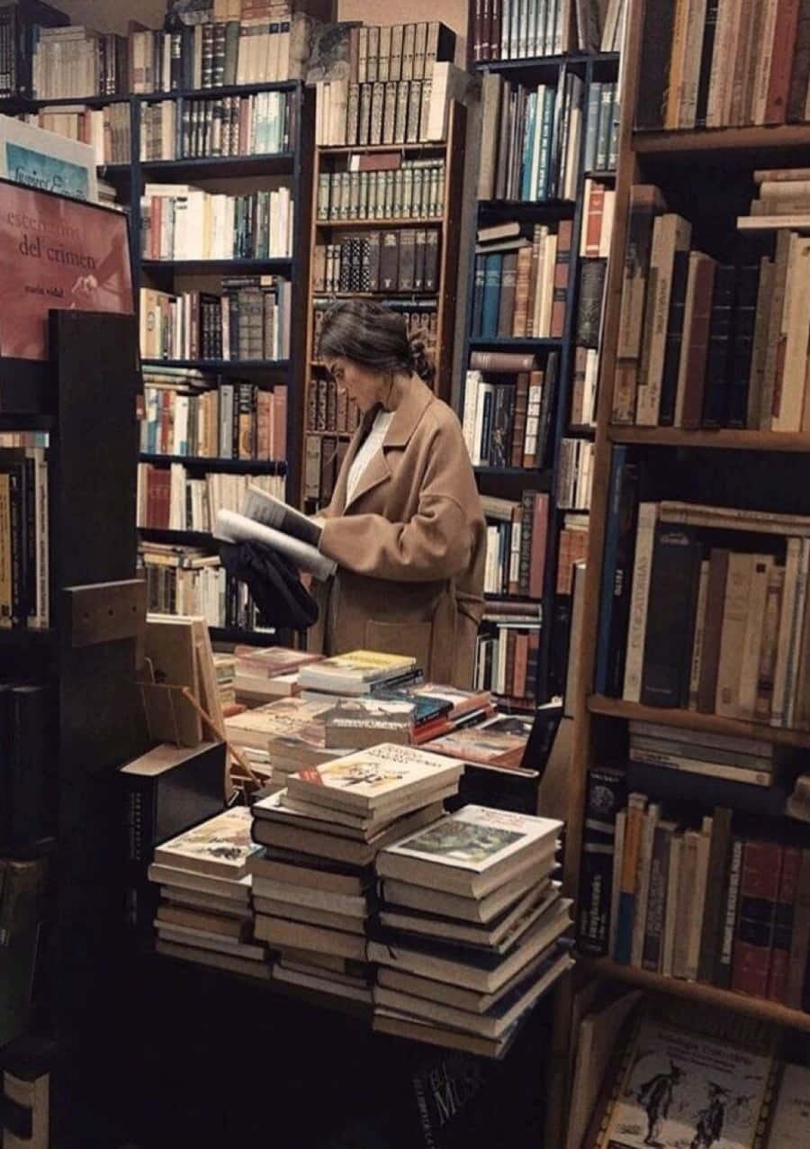圖書館內閱讀的女學生 壁紙 — 一位身穿米色外套的女性在舒適、燈光昏暗的書店中，在堆滿復古書籍的高聳書架間閱讀一本書。