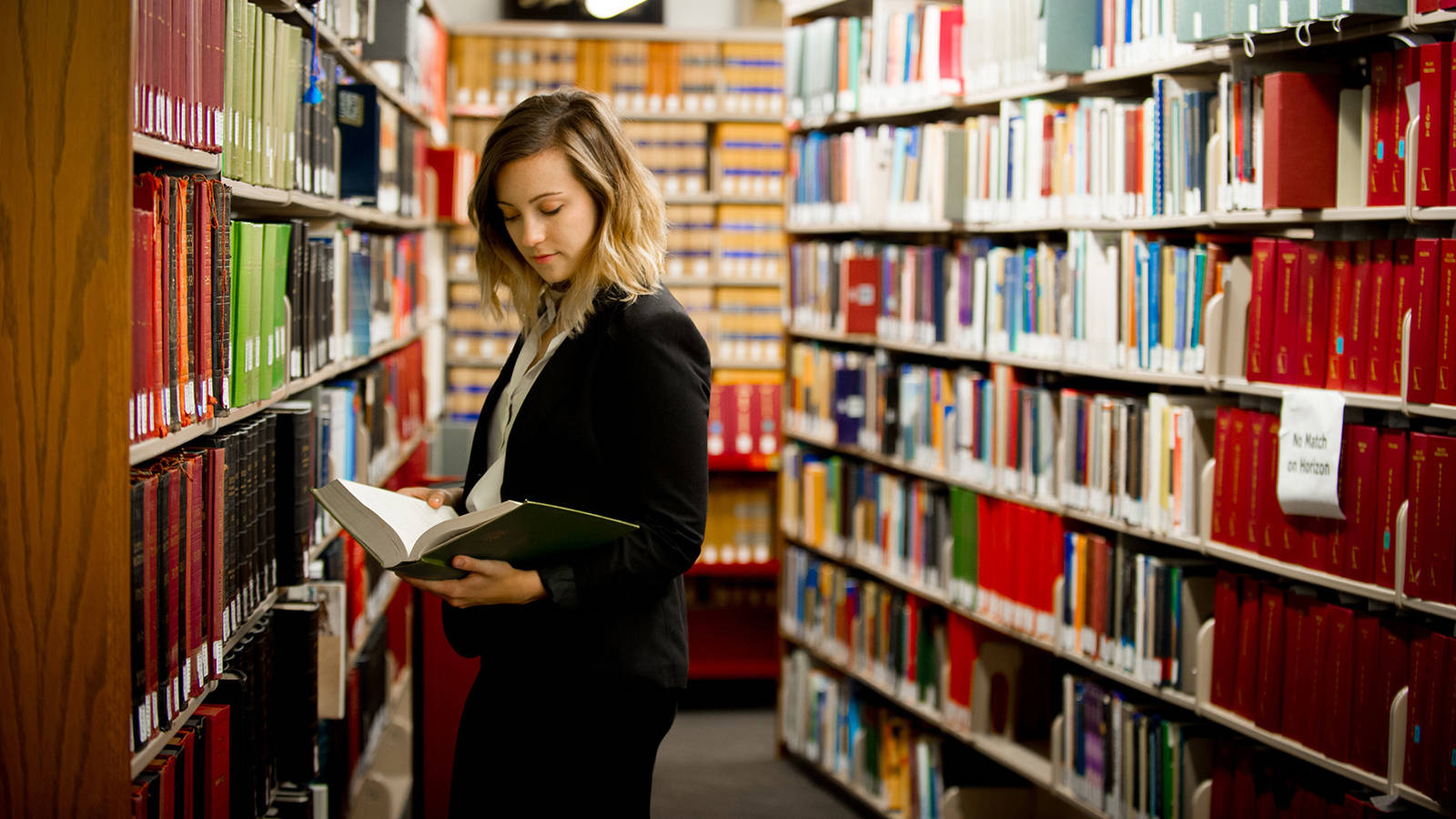 穿著黑色西裝外套的女性在圖書館書架之間閱讀書籍。