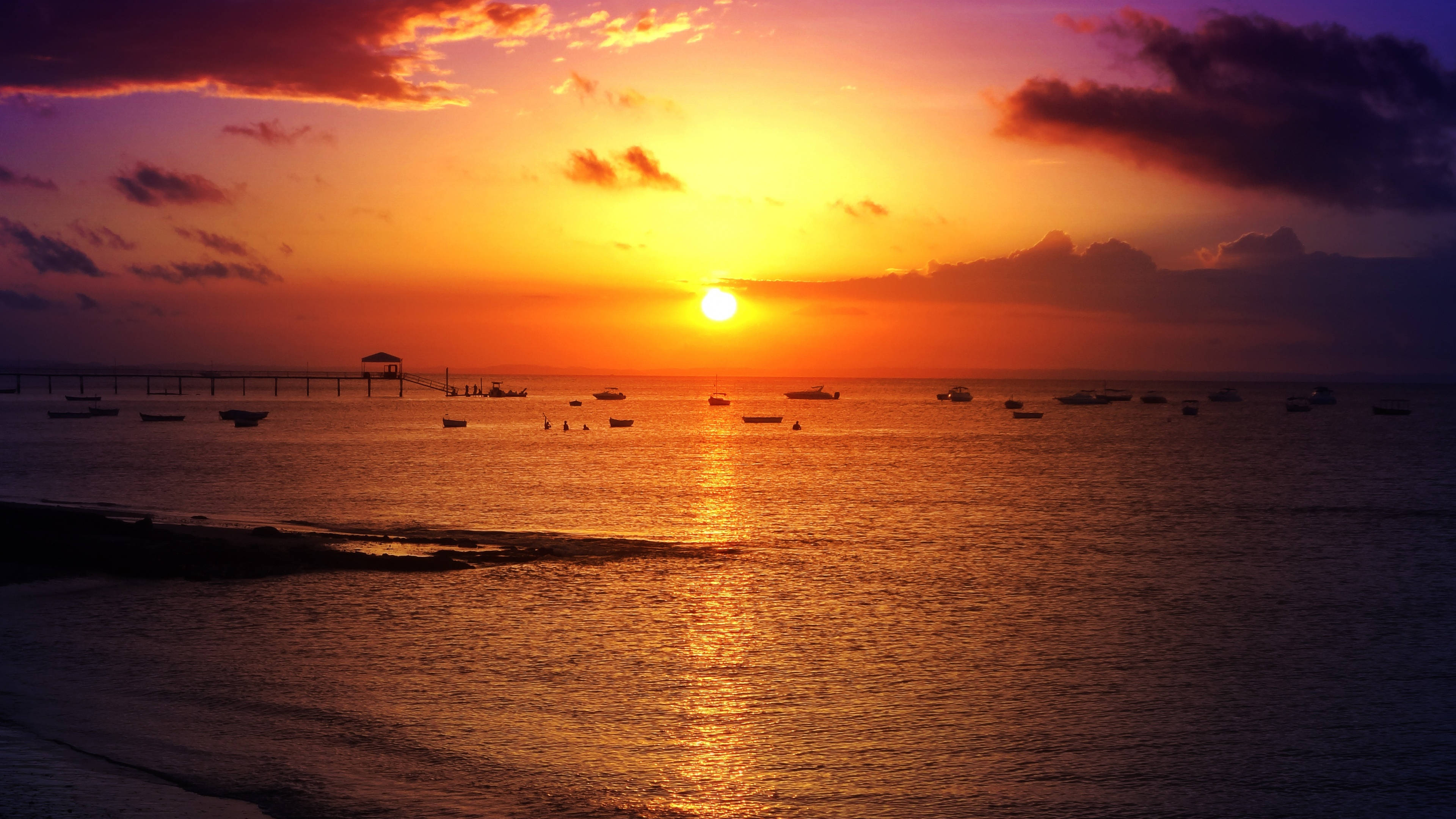 圖瓦利日落天空 壁紙 — 夕陽燦爛，天空染上熾熱的橙色和紫色，船隻在碼頭附近的平靜水面上休憩。