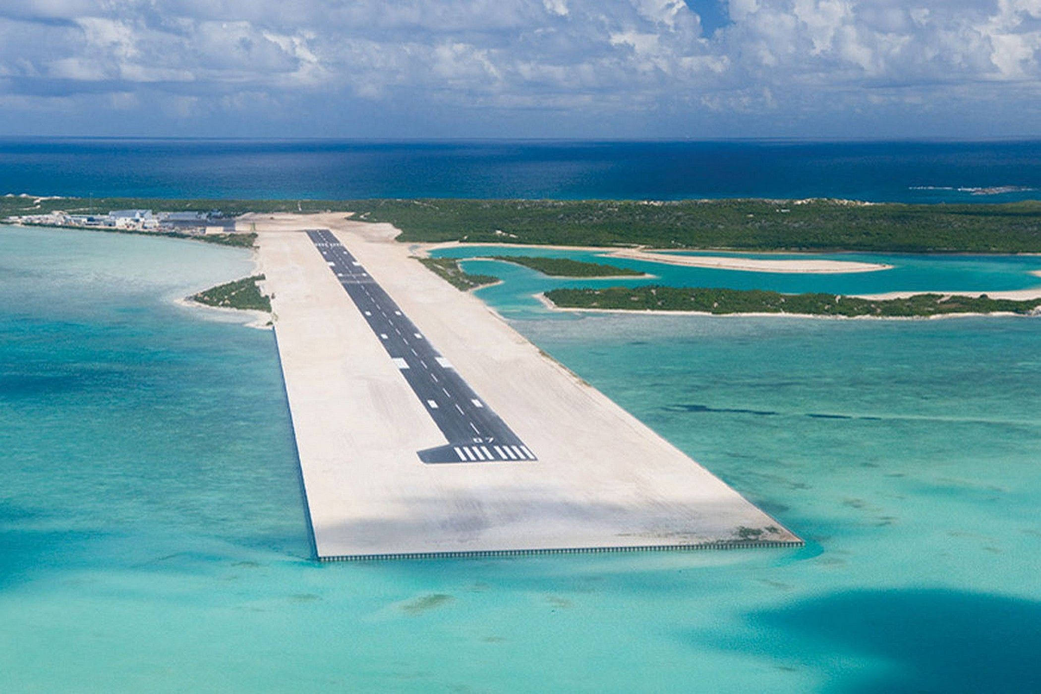 土克島與開基島機場 壁紙 — 從空中俯瞰一條長長的混凝土跑道，延伸在碧綠色的海水上，通往一個鬱鬱蔥蔥的綠色島嶼，天空部分多雲。