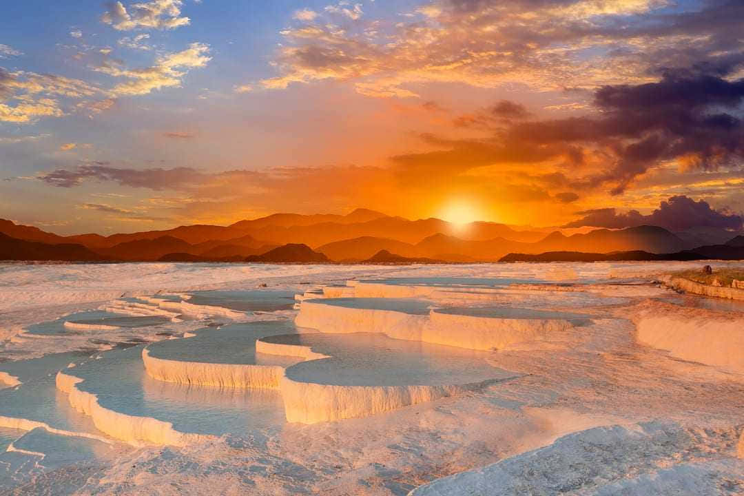 土耳其地中海沿岸的美麗日落 壁紙 — 夕陽將梯田溫泉沐浴在溫暖的橙色光芒中，背景是遠山和戲劇性的雲彩。