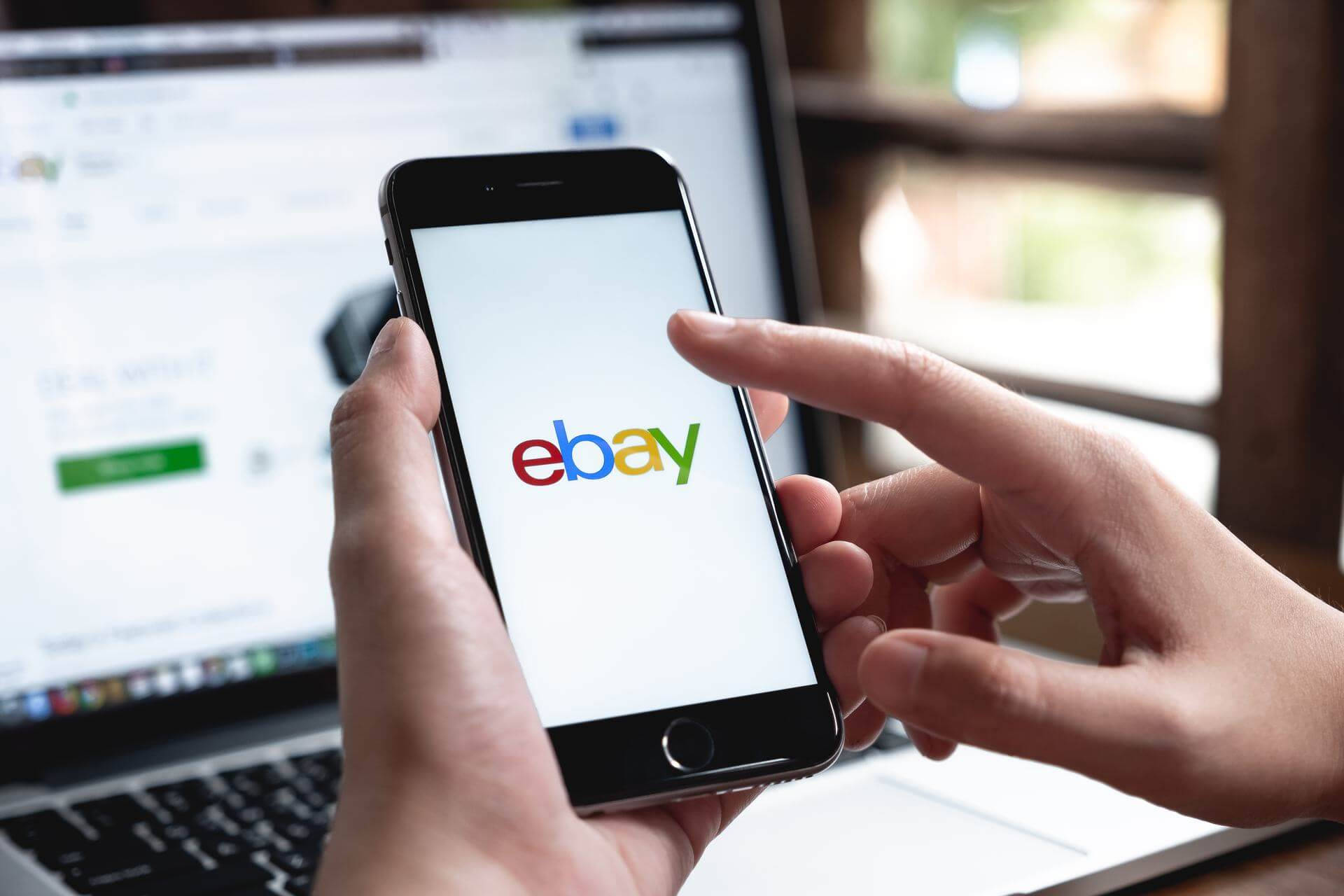 在 eBay 上瀏覽 壁紙 — 雙手拿著顯示eBay標誌的智能手機，背景中可見同一網站的筆記本電腦。