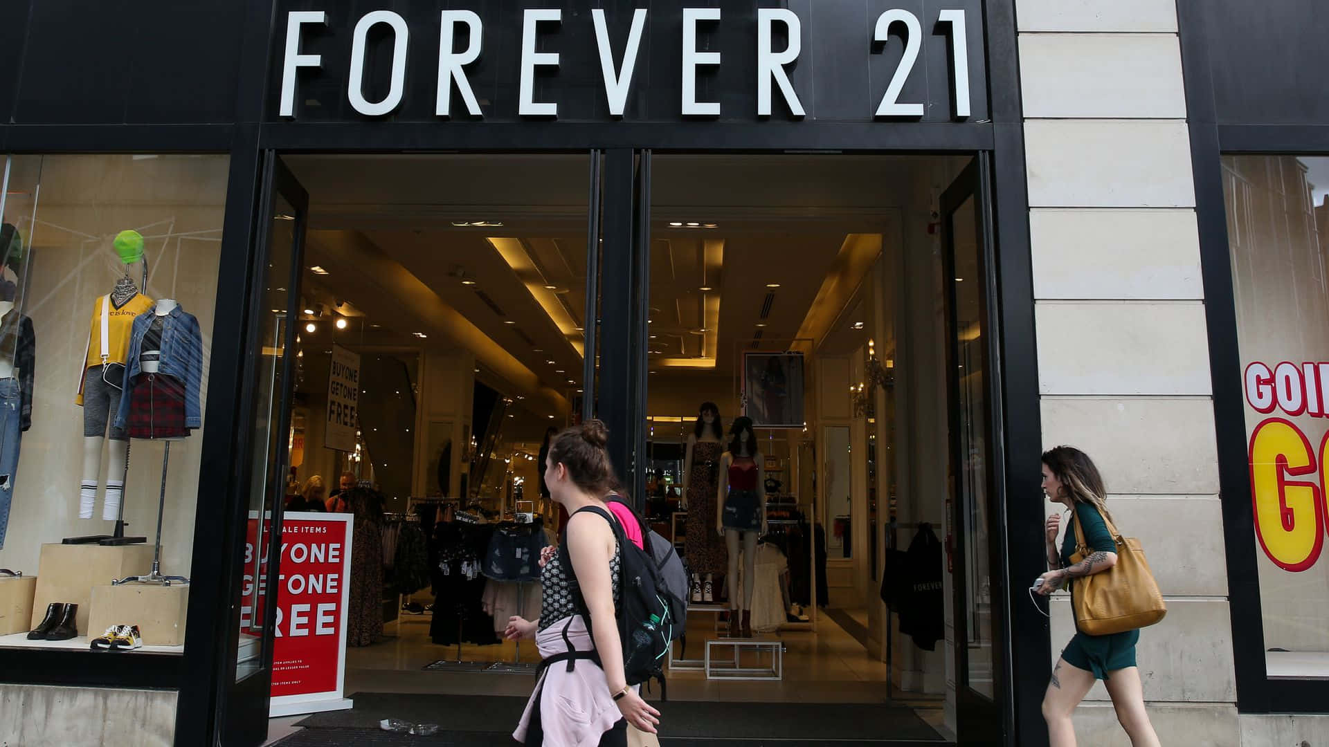 在 Forever 21 找到超值優惠，打造全新衣櫥 壁紙 — 兩位女士走過一間Forever 21商店，店舖有明亮的招牌和櫥窗內的模特兒。