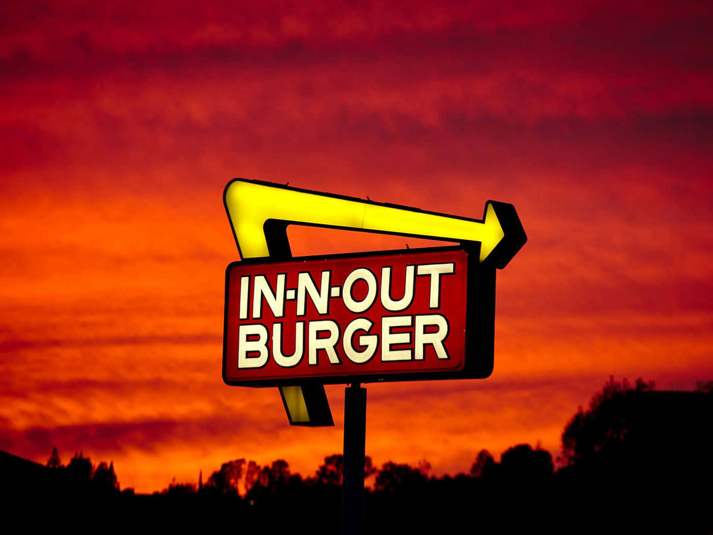一個明亮的黃色箭頭標誌，代表 In-N-Out Burger，映襯著充滿活力的日落天空。