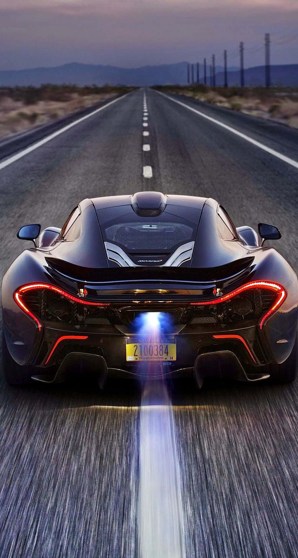 在 iPhone XS Max 上駕駛《Project CARS 2》挑戰極限 壁紙 — 一輛黑色McLaren P1在黃昏時分沿沙漠公路飛馳，帶有發光的紅色尾燈和藍色排氣火焰。