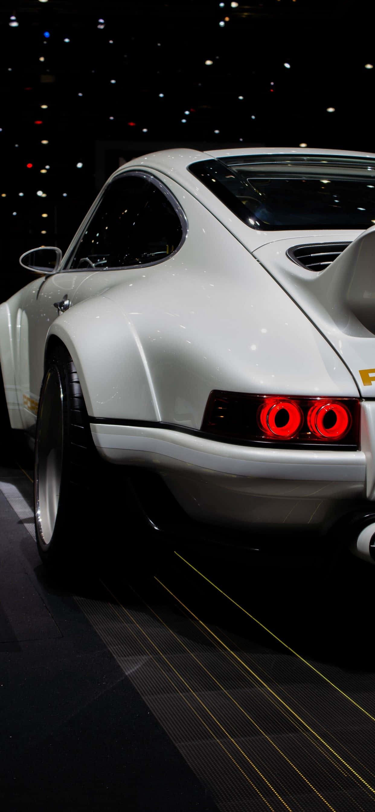 在 iPhone XS Max 上體驗《Project CARS 2》的極速快感 壁紙 — 一架流線型白色經典跑車，帶有發光的紅色尾燈，在黑暗展廳的聚光燈下展示。