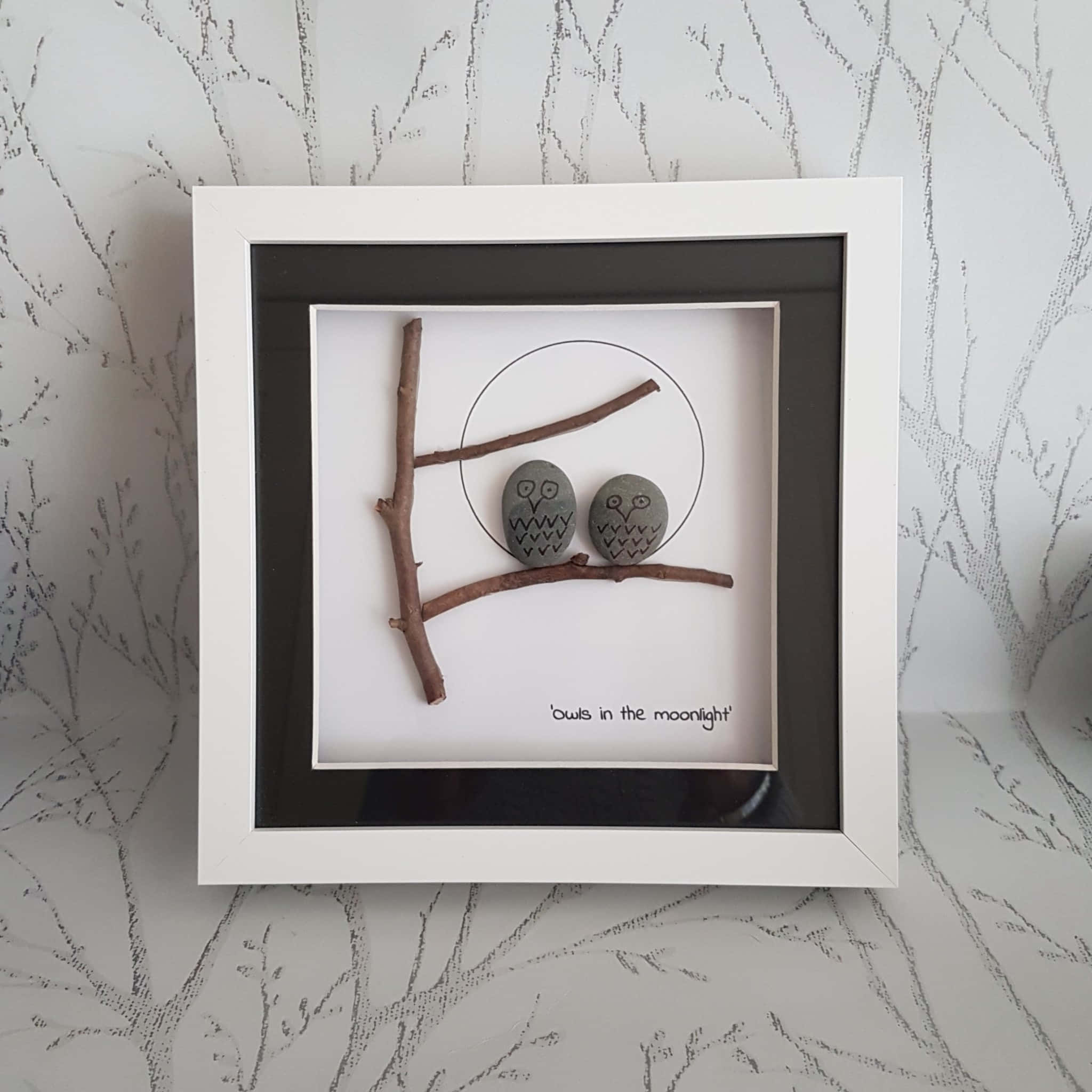 在 Pebble Art 的美中尋得寧靜 壁紙 — 兩隻灰色石頭貓頭鷹坐在樹枝上，下方有繪製的月亮，置於白色陰影框中，背景為樹紋牆壁。