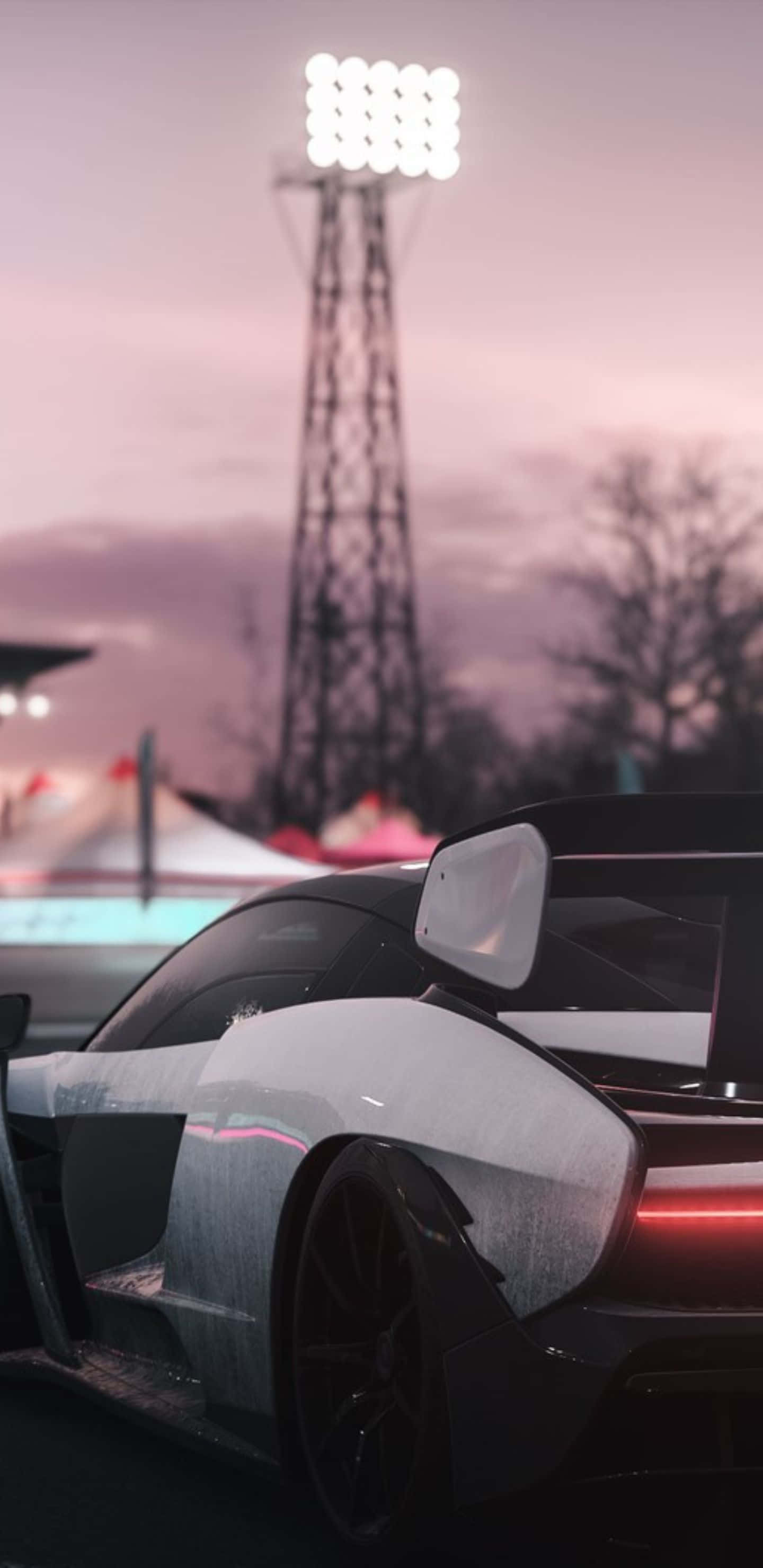 在 Pixel 3XL 和 Forza Horizon 4 上體驗震撼視覺與遊戲樂趣 壁紙 — 一架時尚的黑色賽車，帶有發光的紅色尾燈，停泊在暮色中的體育場燈塔下。