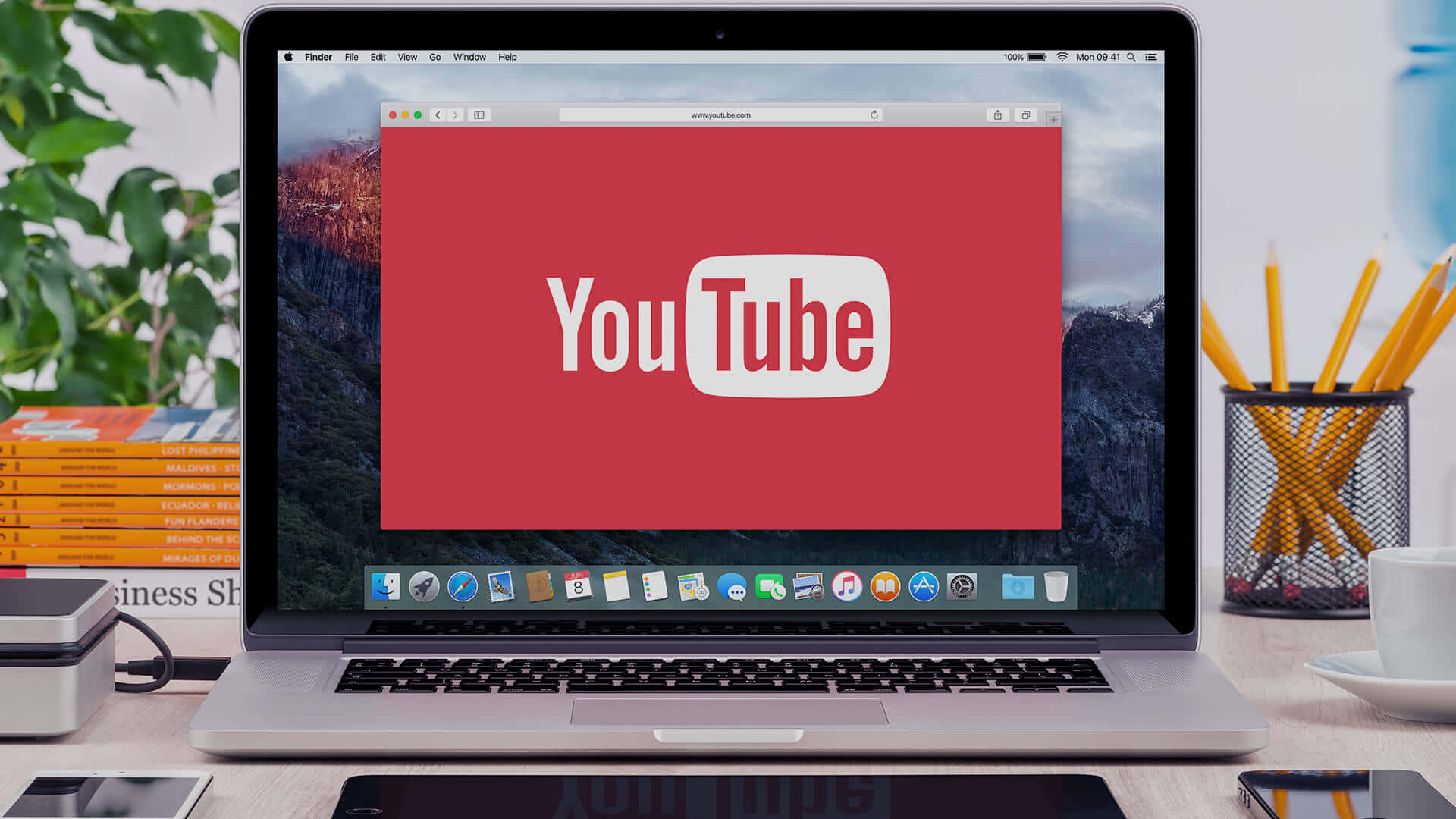 在 YouTube 上分享更多內容 壁紙 — 一部銀色手提電腦在紅色屏幕上顯示YouTube標誌，周圍有山脈桌面背景、辦公用品和植物。