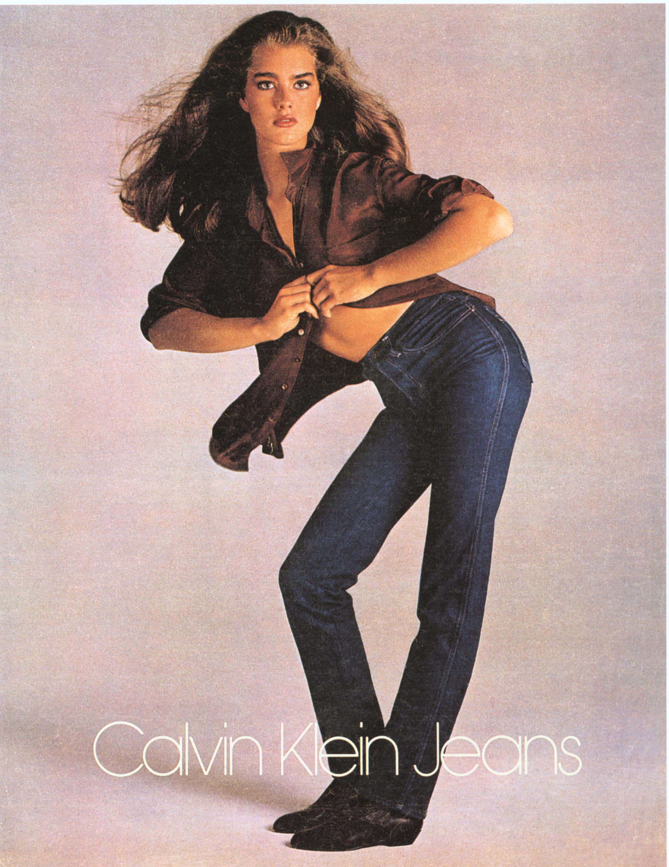 在Calvin Klein的經典優雅中找到時尚自信 壁紙 — 一位擁有飄逸深色頭髮的女性，身穿藍色牛仔褲和棕色襯衫，在柔和的灰色工作室背景前自信地擺姿勢。