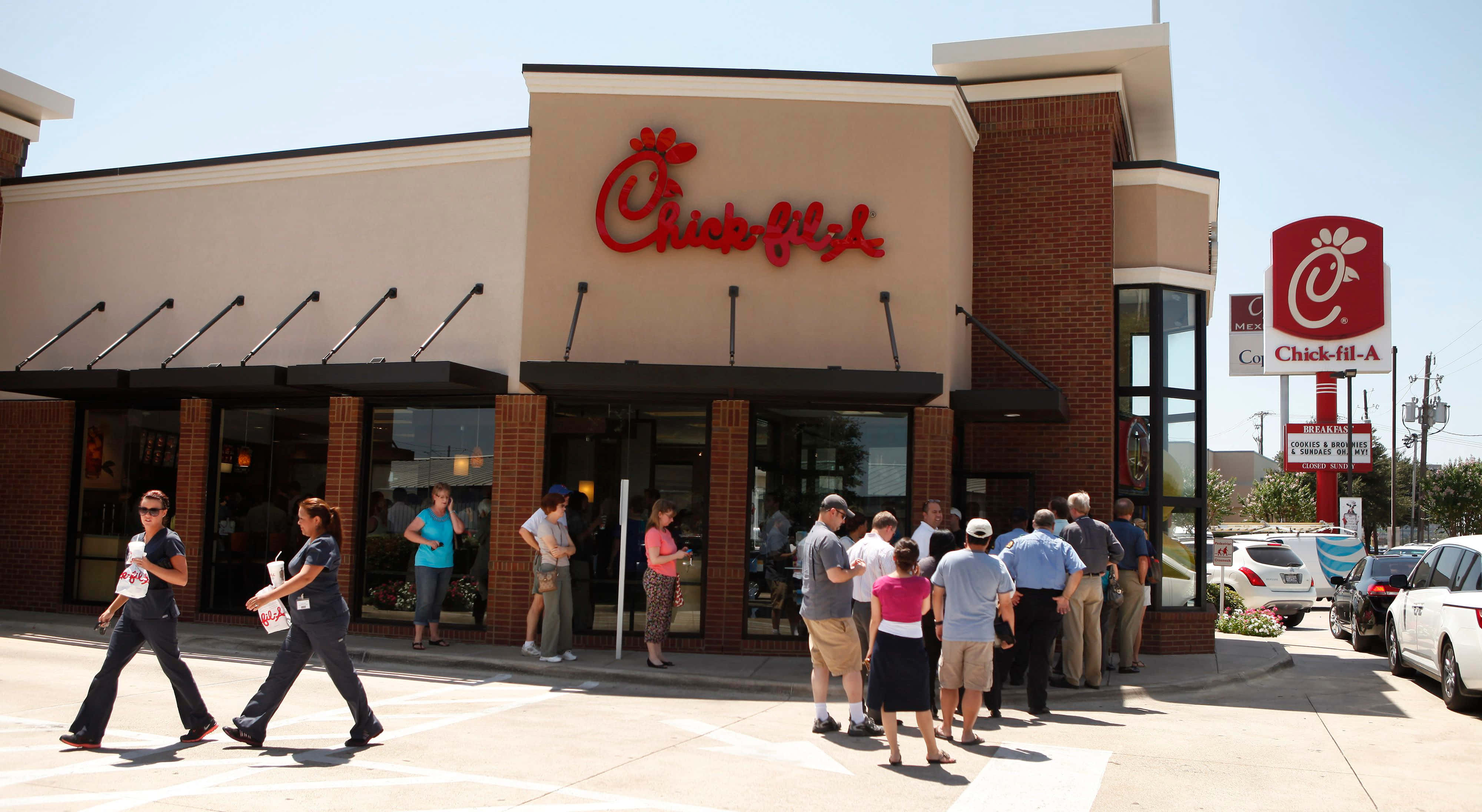 在Chick-fil-A享受美味餐點！ 壁紙 — 顧客在明亮的日光下在Chick-fil-A餐廳外排隊，附近有停泊的車輛。