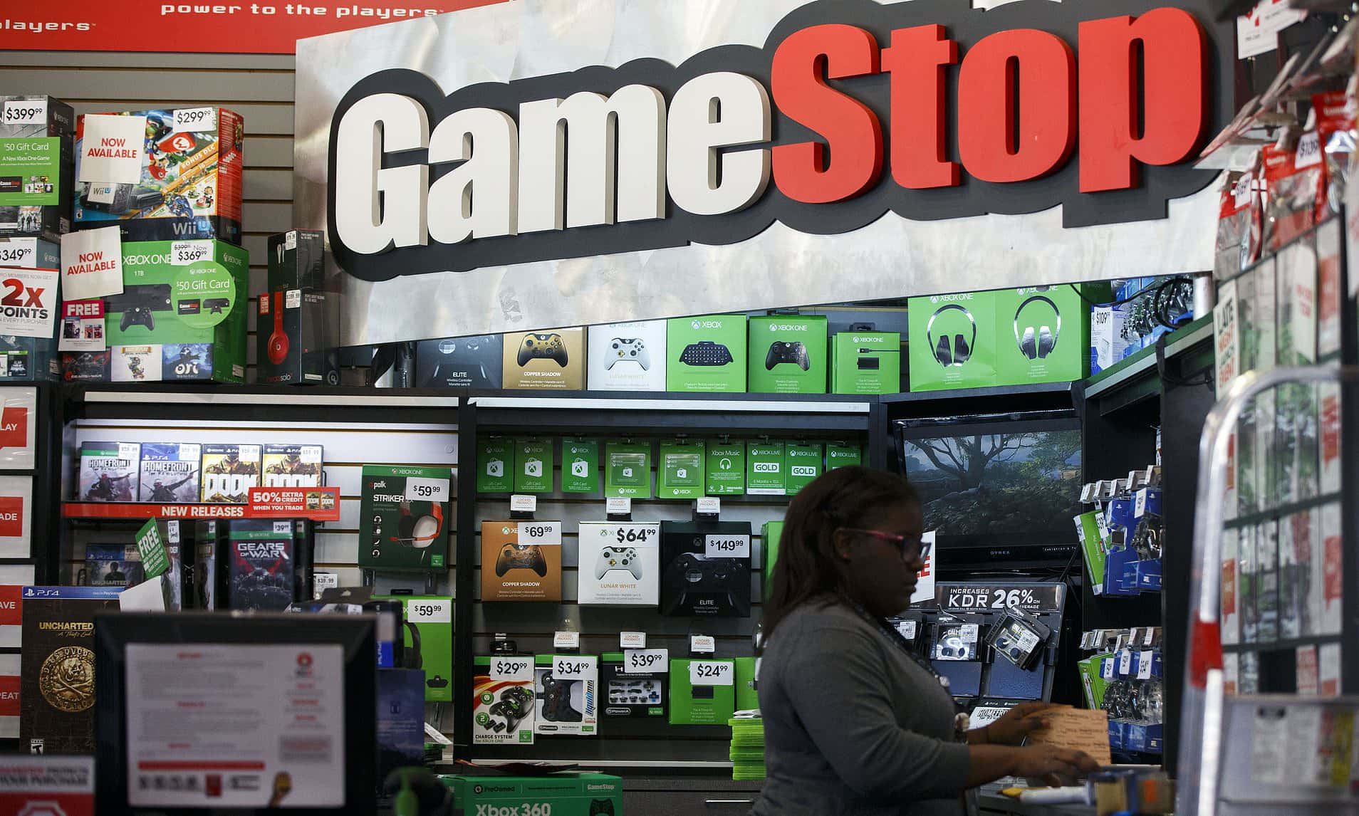 在GameStop找到你的下一款最愛遊戲 壁紙 — 一名女性在GameStop零售店內明亮的店舖燈光下瀏覽電子遊戲商品。