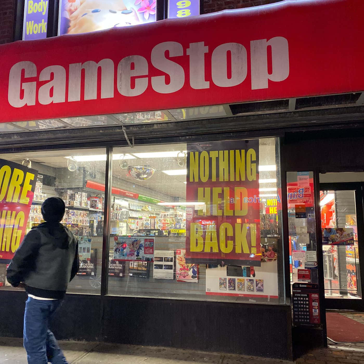 在GameStop找到遊戲天堂 壁紙 — 夜晚，一個人走過一間燈火通明的GameStop商店，窗戶上有醒目的招牌和促銷海報。