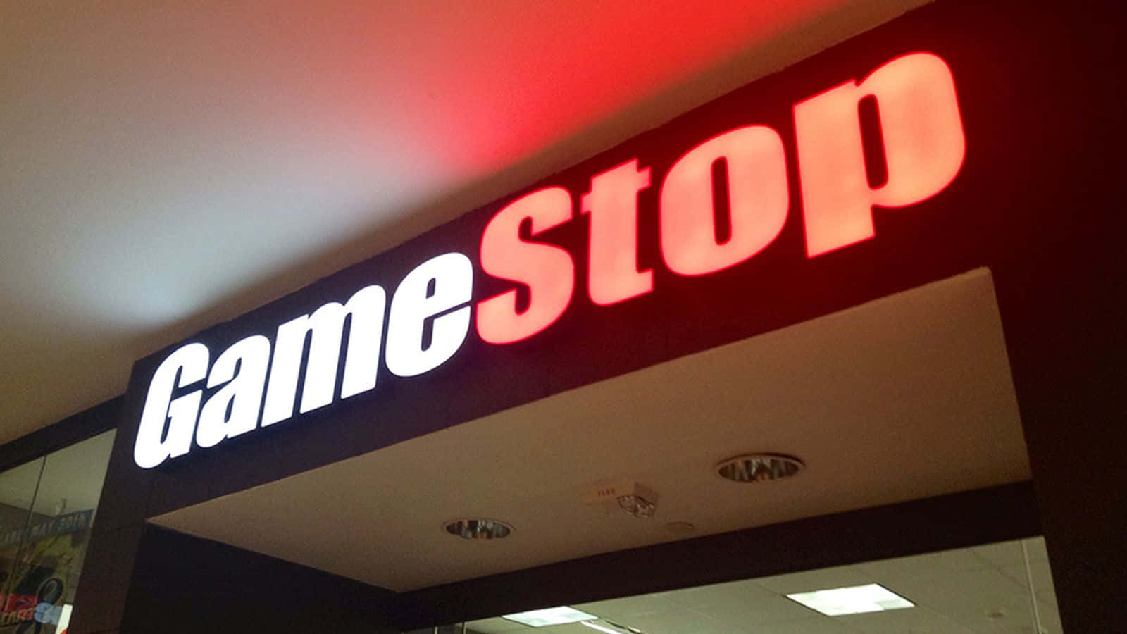 在GameStop提升您的遊戲時光 壁紙 — 低角度拍攝一個發光紅白色GameStop商店標誌，背景為黑暗天花板及嵌入式燈光。