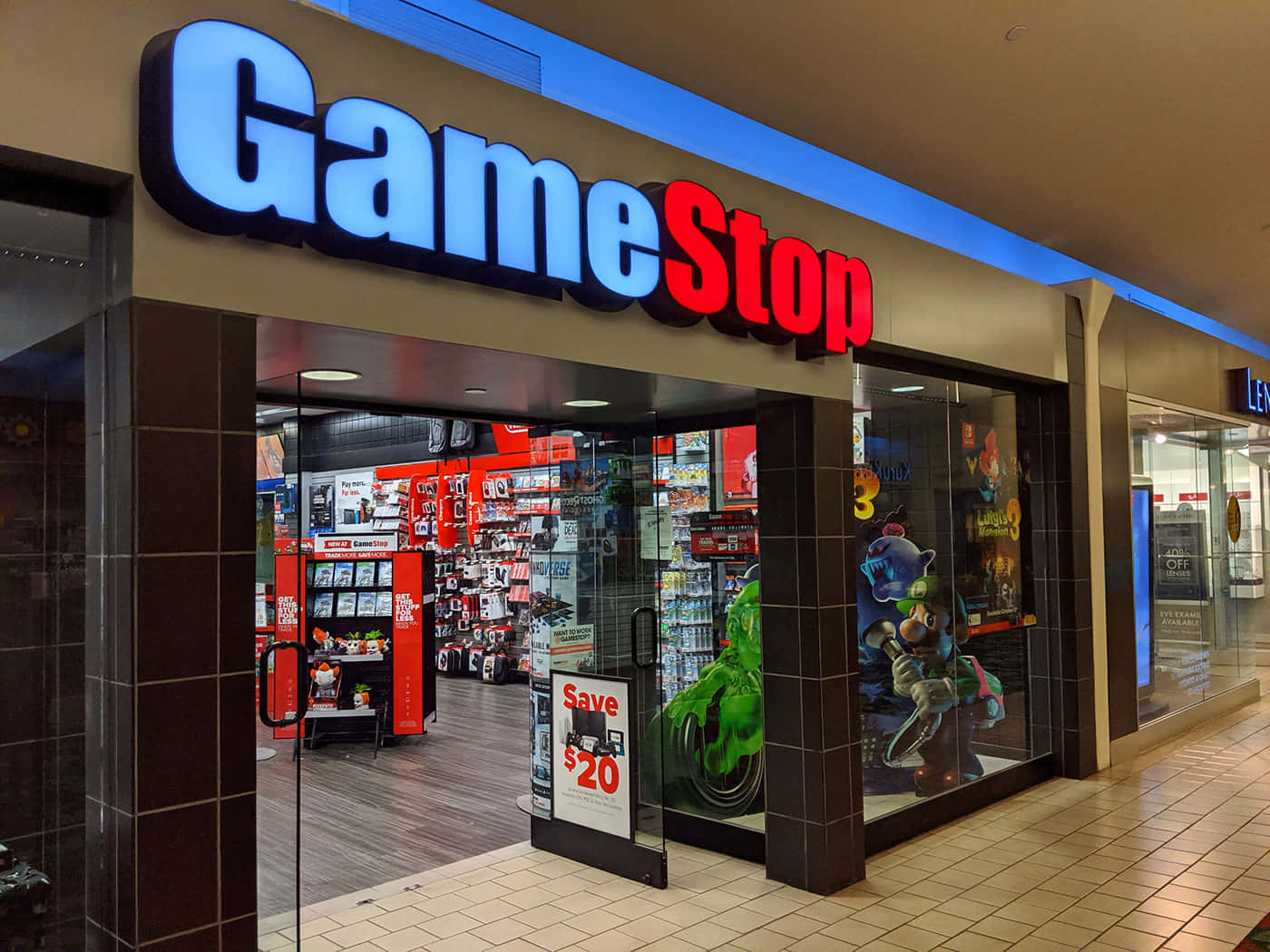 在GameStop準備你下一款最愛遊戲！ 壁紙 — 光線明亮的GameStop店舖正面，玻璃門窗展示商場內色彩繽紛的電玩商品和角色立牌。