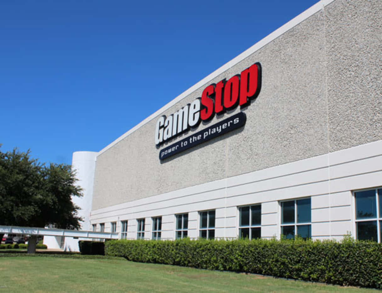 在GameStop玩您最愛的遊戲 壁紙 — GameStop 商店建築物，在晴朗藍天下，前景有綠色籬笆和樹木。