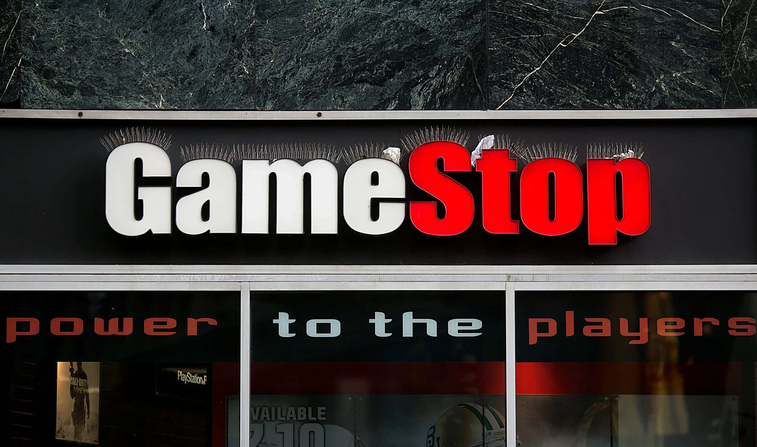 在Gamestop購買您最愛的遊戲！ 壁紙 — GameStop 店舖招牌，以粗體紅白字體顯示，背景為深色紋理牆壁。