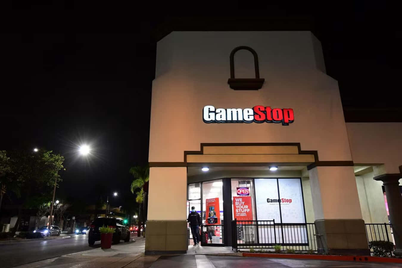 在Gamestop購買新舊遊戲！ 壁紙 — 夜晚明亮照明的GameStop商店，有人在發光招牌下進入，街燈照亮行人路。