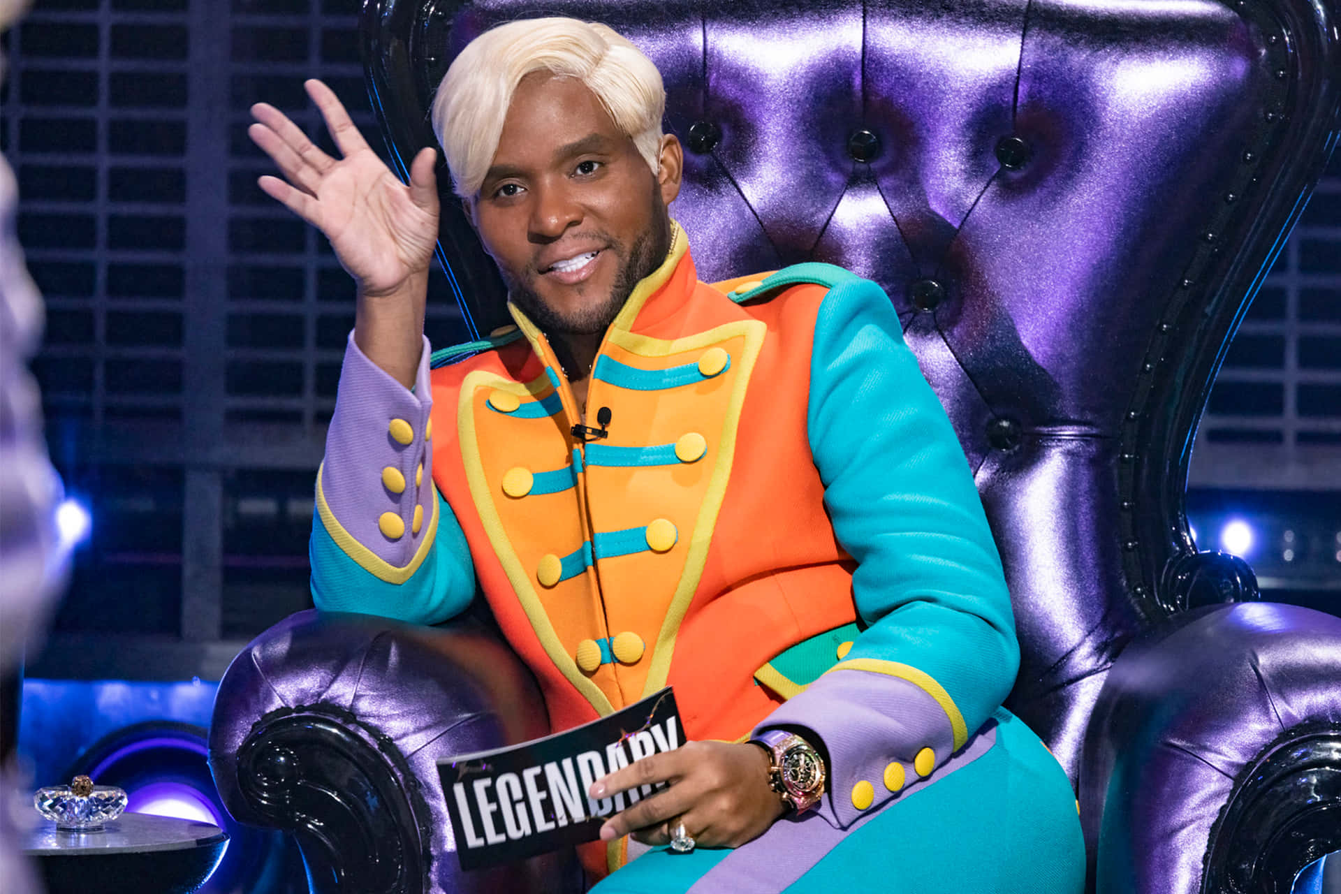 穿著色彩繽紛服裝的男子從紫色王座椅上揮手，手持「LEGENDARY」標誌。