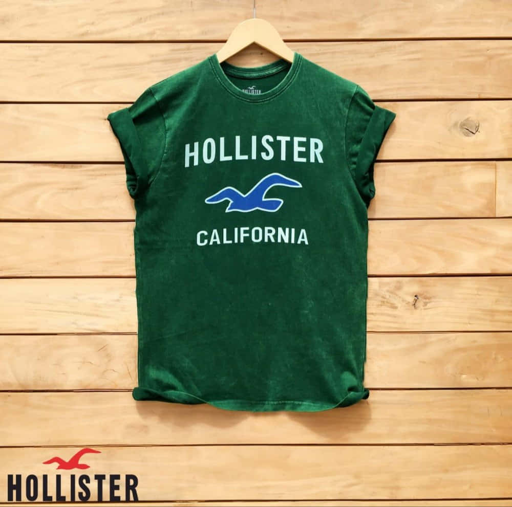 綠色Hollister California T恤掛在木衣架上，背景為木牆。
