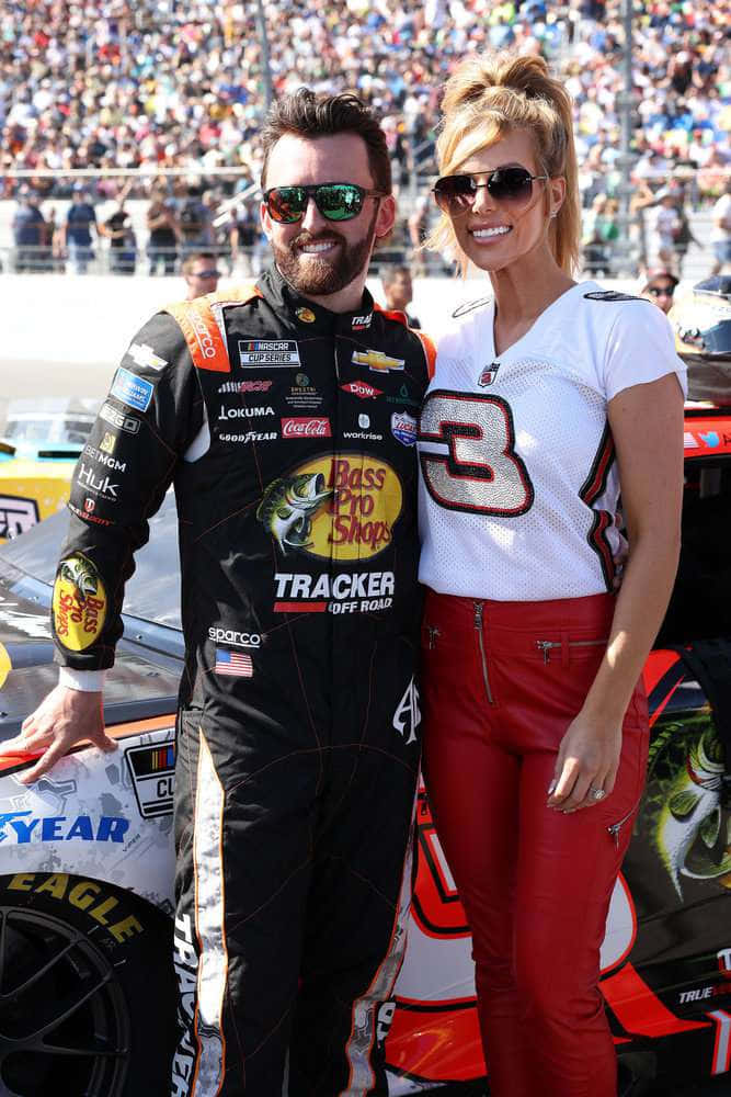 在NASCAR賽事中突破極速 壁紙 — 一位微笑的NASCAR車手和一位金髮賽車女郎在陽光明媚的賽道上，於一輛賽車旁擺姿勢，賽道擠滿了觀眾。
