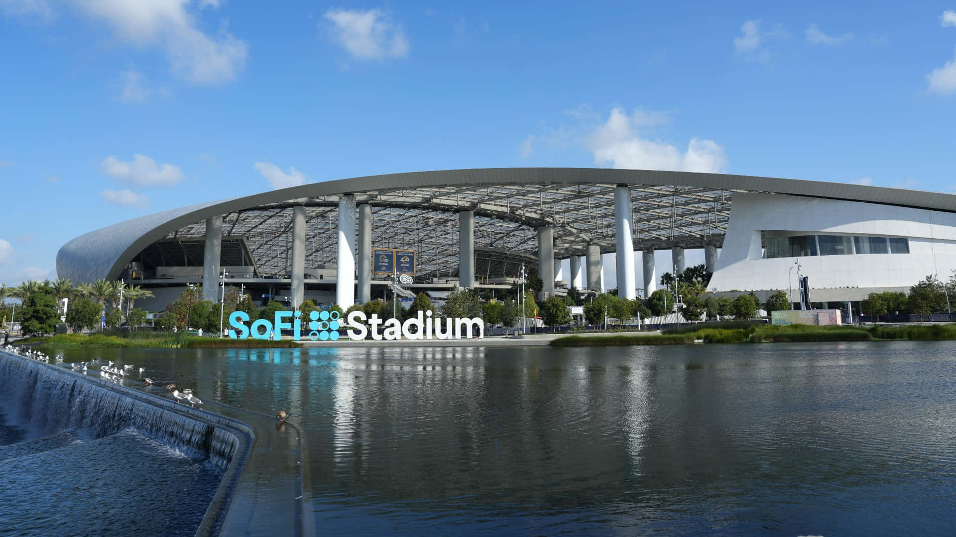在SoFi體育館體驗比賽的刺激 壁紙 — SoFi 體育場在明亮的藍天白雲下閃耀，其現代建築倒映在平靜的水面上，鳥兒聚集在小瀑布附近。