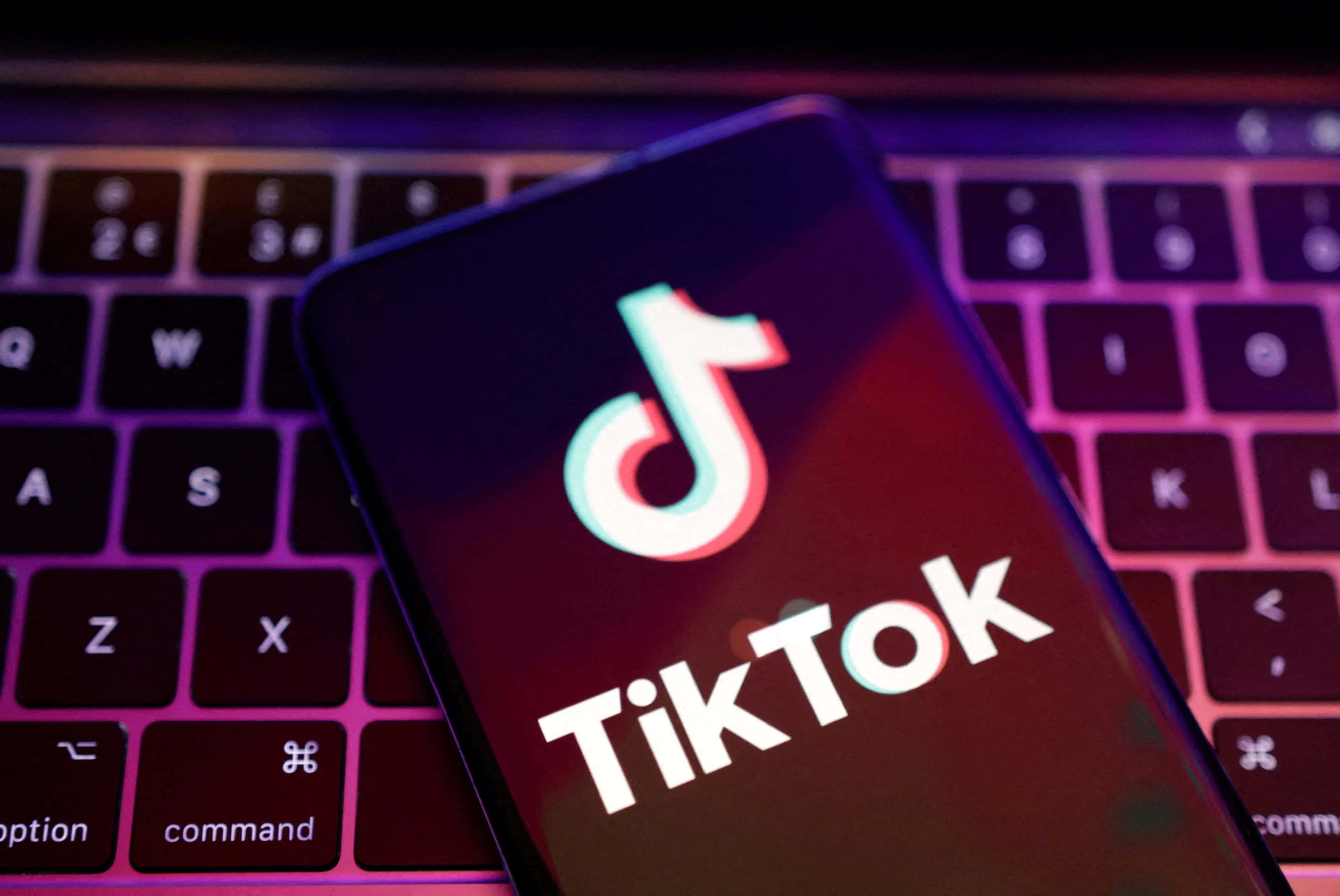 在TikTok上盡情享受夜晚！ 壁紙 — 一部顯示TikTok標誌的智能手機放在一個沐浴在紫色和粉紅色霓虹燈光下的鍵盤上。