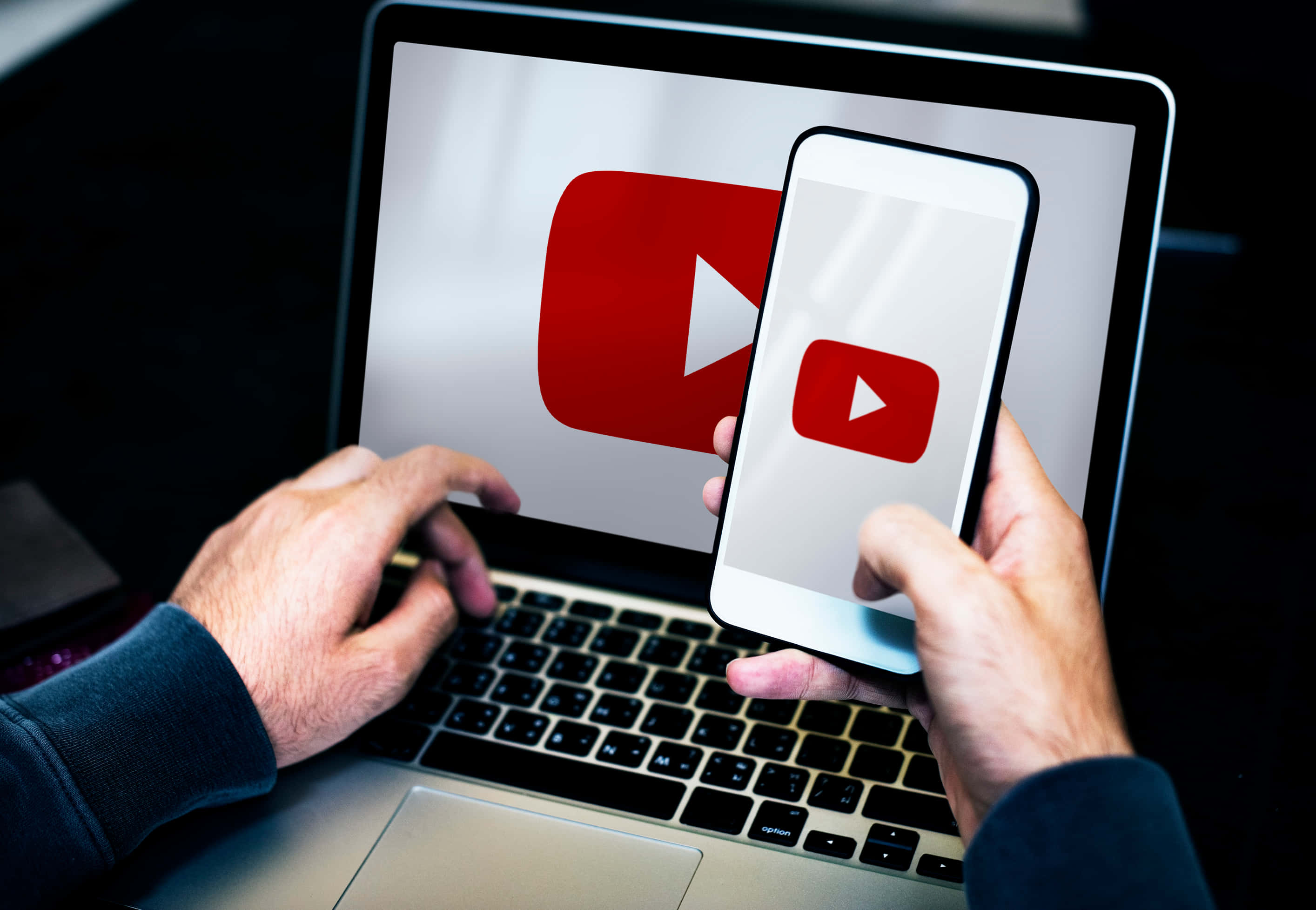 在YouTube上發揮創意！ 壁紙 — 雙手握住智能手機和手提電腦，兩者都在昏暗的房間中顯示YouTube標誌。
