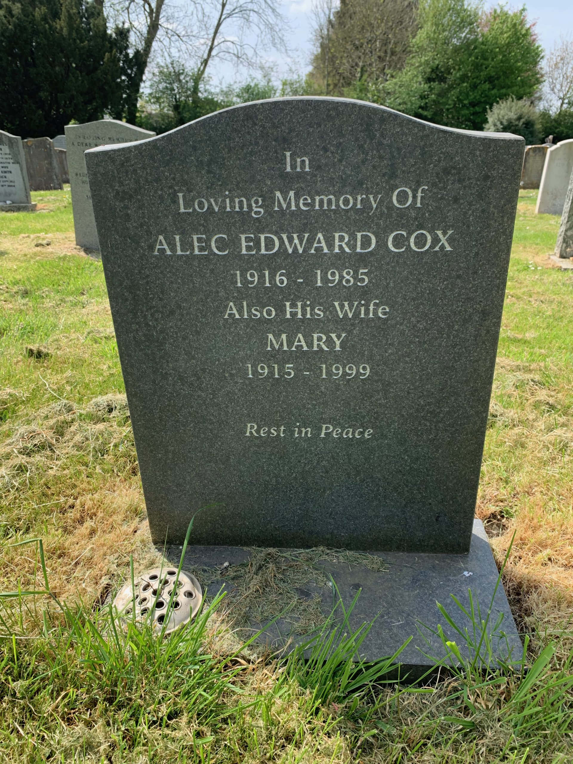 Gravestone 桌布 壁紙 — 在一個長滿草的墓園裡，一塊深灰色的墓碑紀念著Alec Edward Cox和他的妻子Mary，上面刻有日期和「安息」字樣，在明亮的日光下。