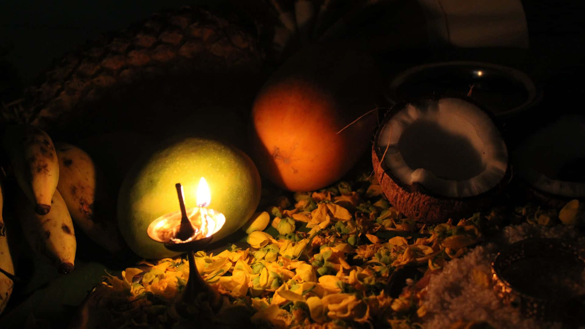 Vishu 桌布 壁紙 — 在一個黑暗的熱帶靜物畫中，一支溫暖的蠟燭在芒果上搖曳，周圍環繞著椰子、香蕉和黃色花瓣。