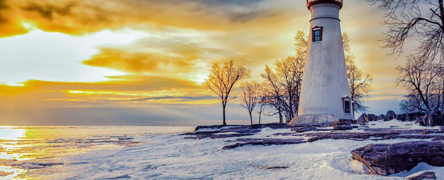 雪岸上的白色燈塔，金色夕陽，光禿樹木和冰冷水面。