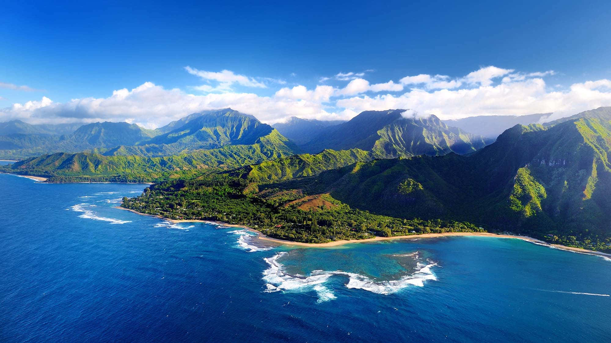 在加勒比海享受悠閒假期 壁紙 — 從高空俯瞰，鬱鬱蔥蔥的青山與碧綠海洋交匯，白色波浪拍打沙灘，天空湛藍明亮。