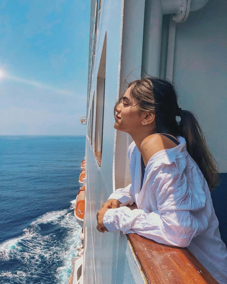 在嘉年華 Vista 船上體驗 Cruise-a-Palooza 壁紙 — 一位身穿白襯衫的年輕女子靠在郵輪的欄杆上，微笑著凝視著燦爛陽光下無盡的藍色海洋。