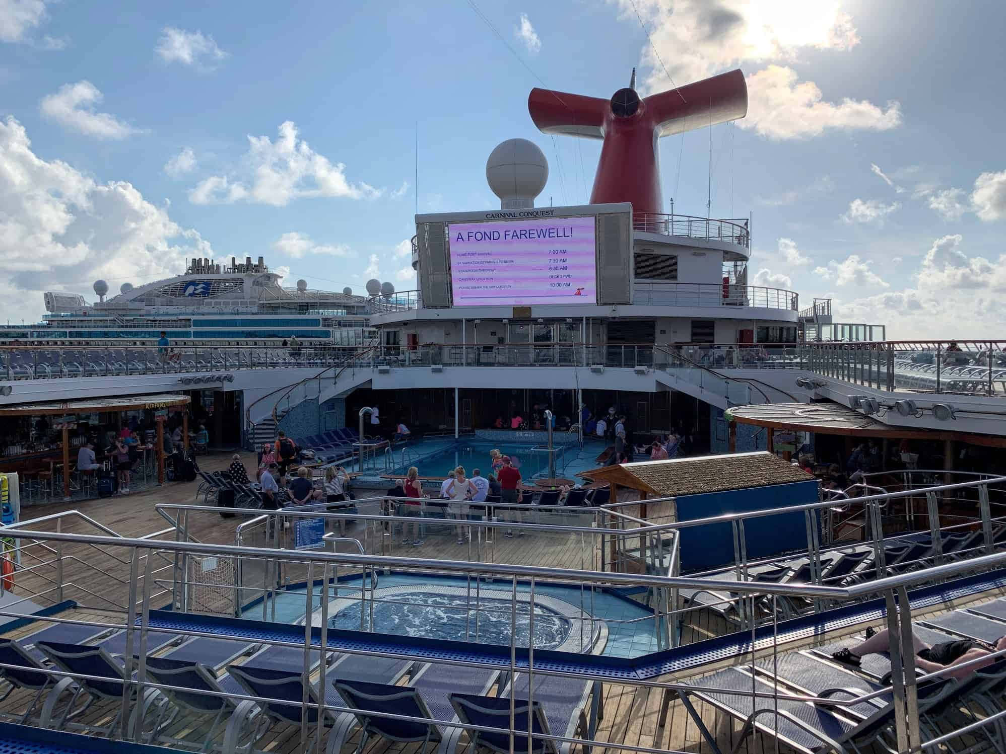 在嘉年華征服號上欣賞加勒比海全景 壁紙 — 在藍天下，Carnival Conquest 遊輪甲板上的一個陽光明媚的日子，設有游泳池、躺椅和告別標誌。