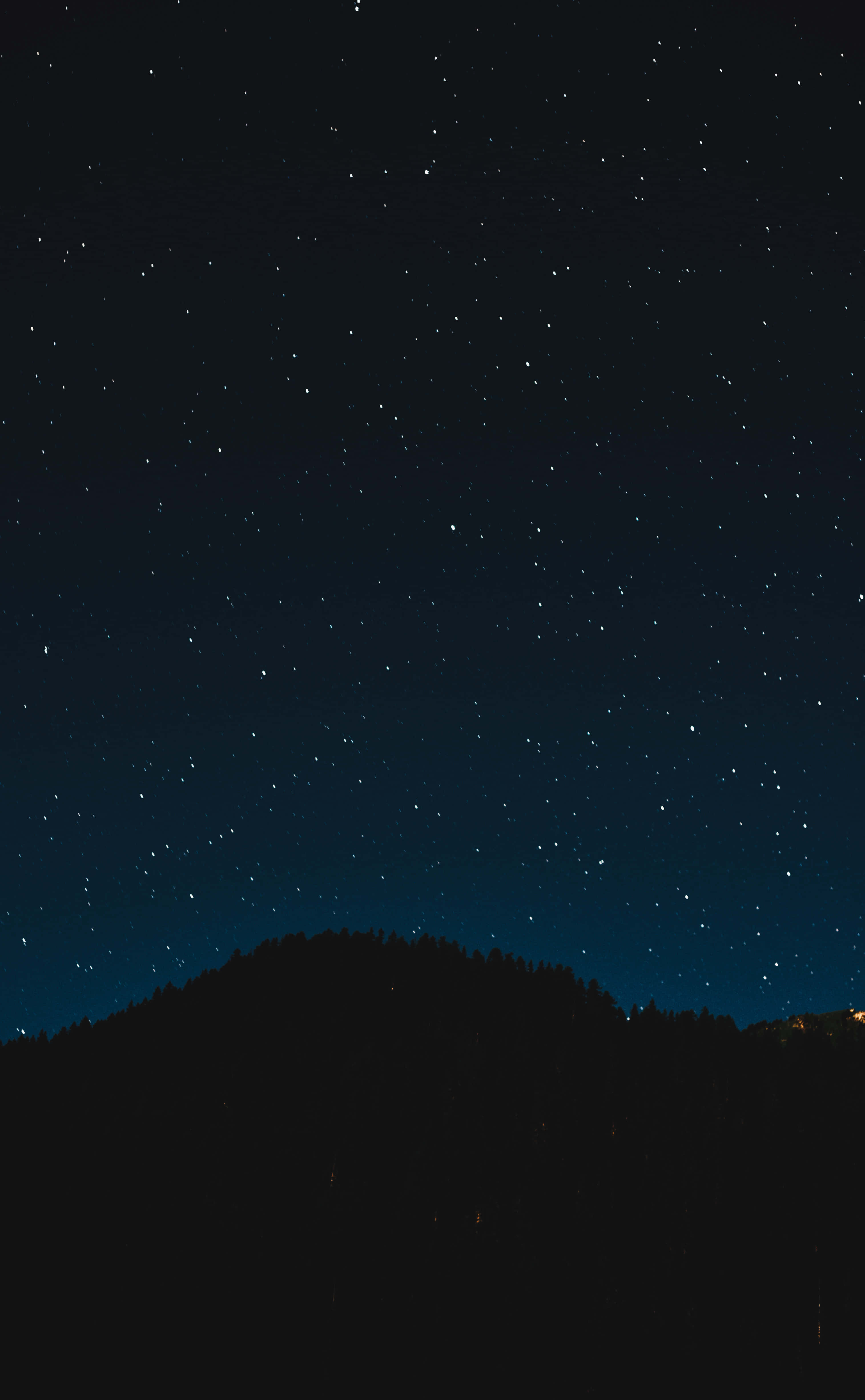 在壯麗夜空下仰望星空 壁紙 — 繁星點點的夜空在黑暗的森林山丘上方閃耀，遠方的燈光在地平線上閃爍。