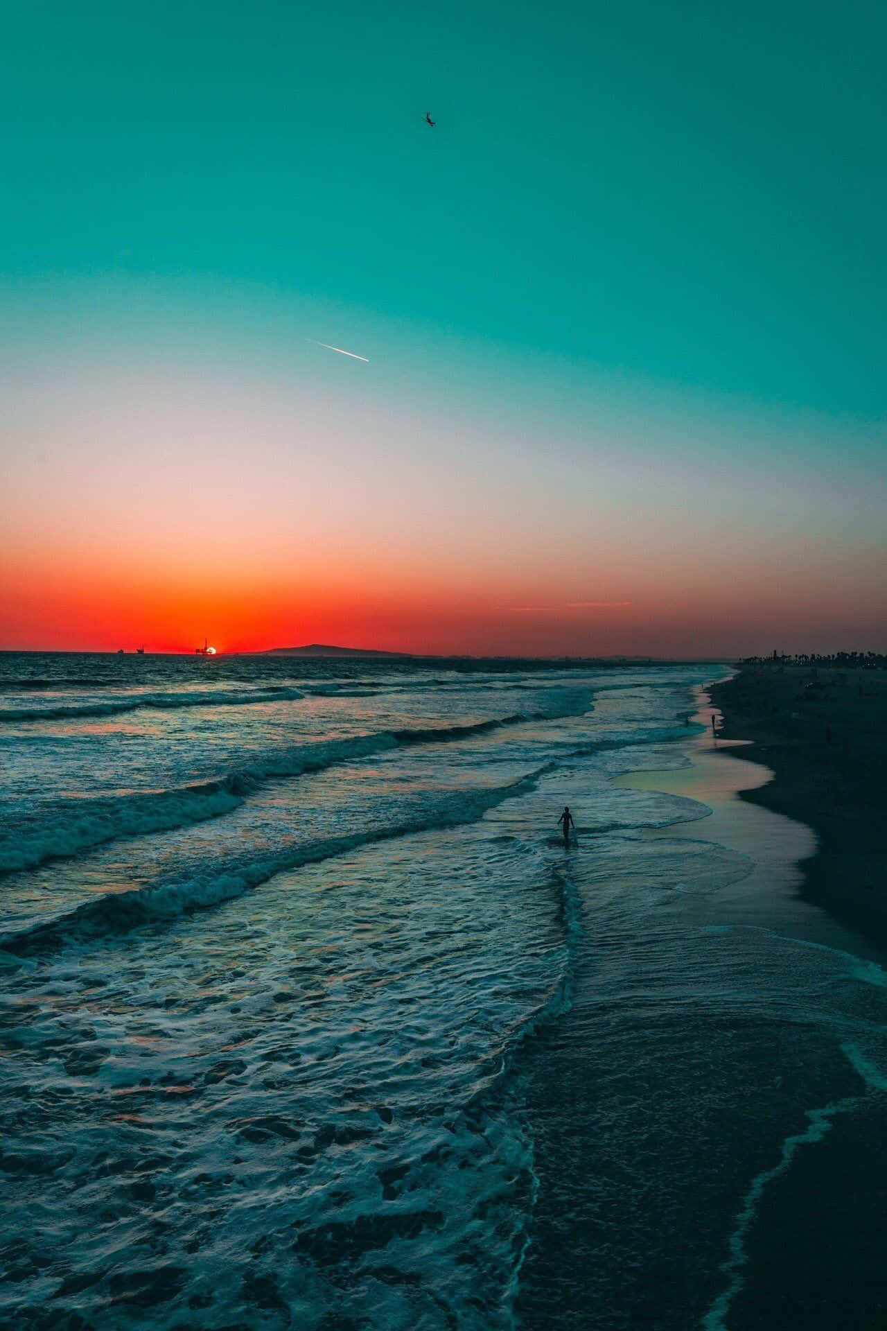 海灘日落 桌布 壁紙 — 在夕陽西下時，一個孤獨的身影沿著海灘行走，海浪在充滿活力的橙色和藍綠色天空下翻滾。