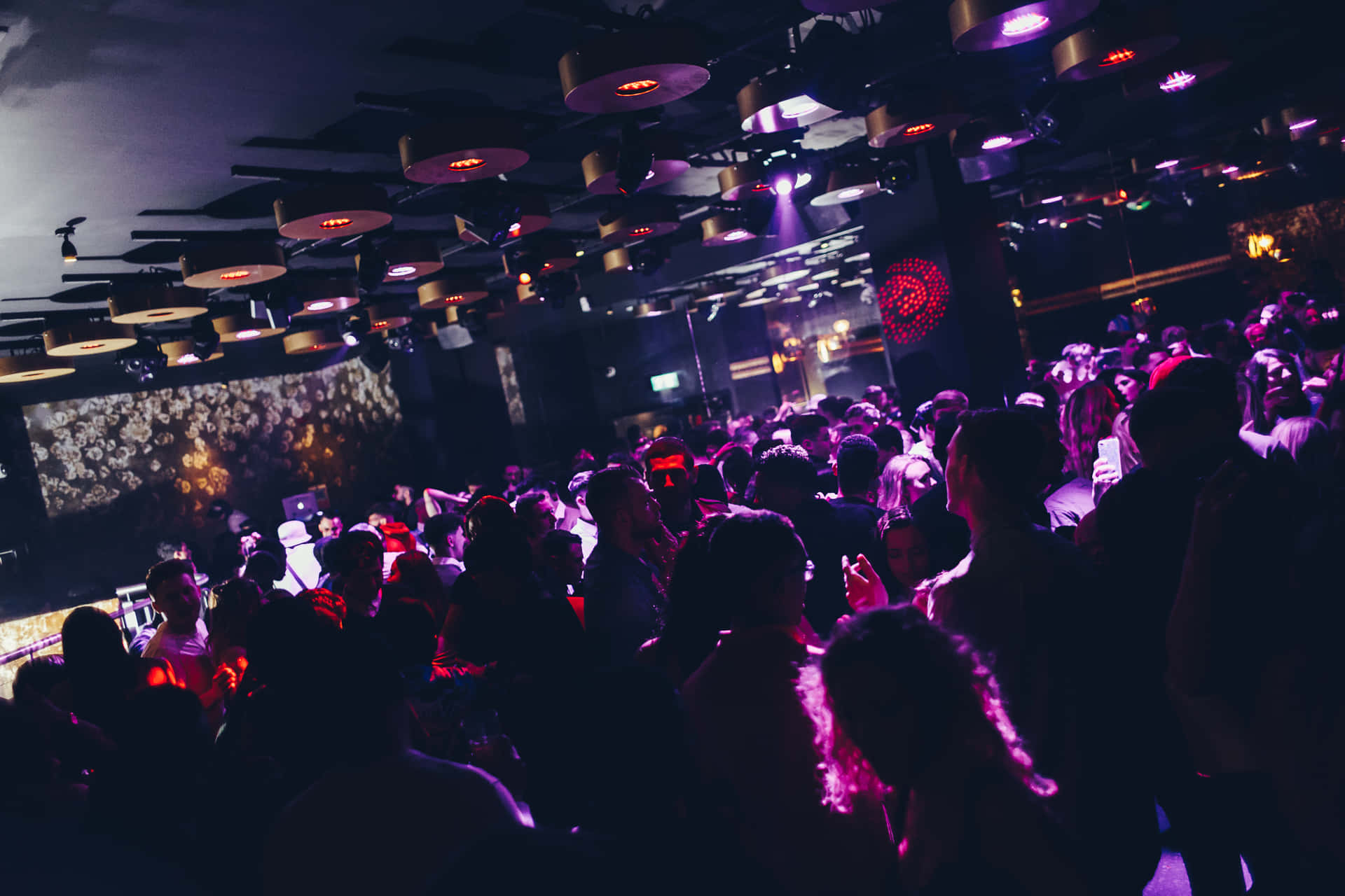在夜店釋放自我，擺脫日常壓力 壁紙 — 人群在昏暗的夜店中，於脈動的紫色燈光、天花風扇和發光舞台裝飾下跳舞。