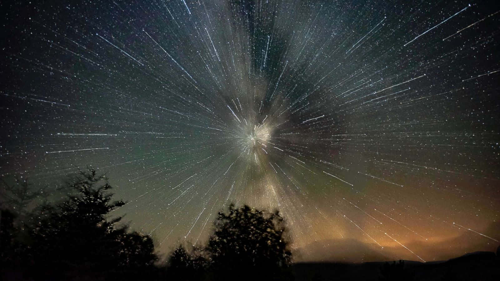 在夜空中飛行的發光流星 壁紙 — 星軌從黑暗夜空中的一個明亮天體點放射而出，上方是剪影般的樹木。