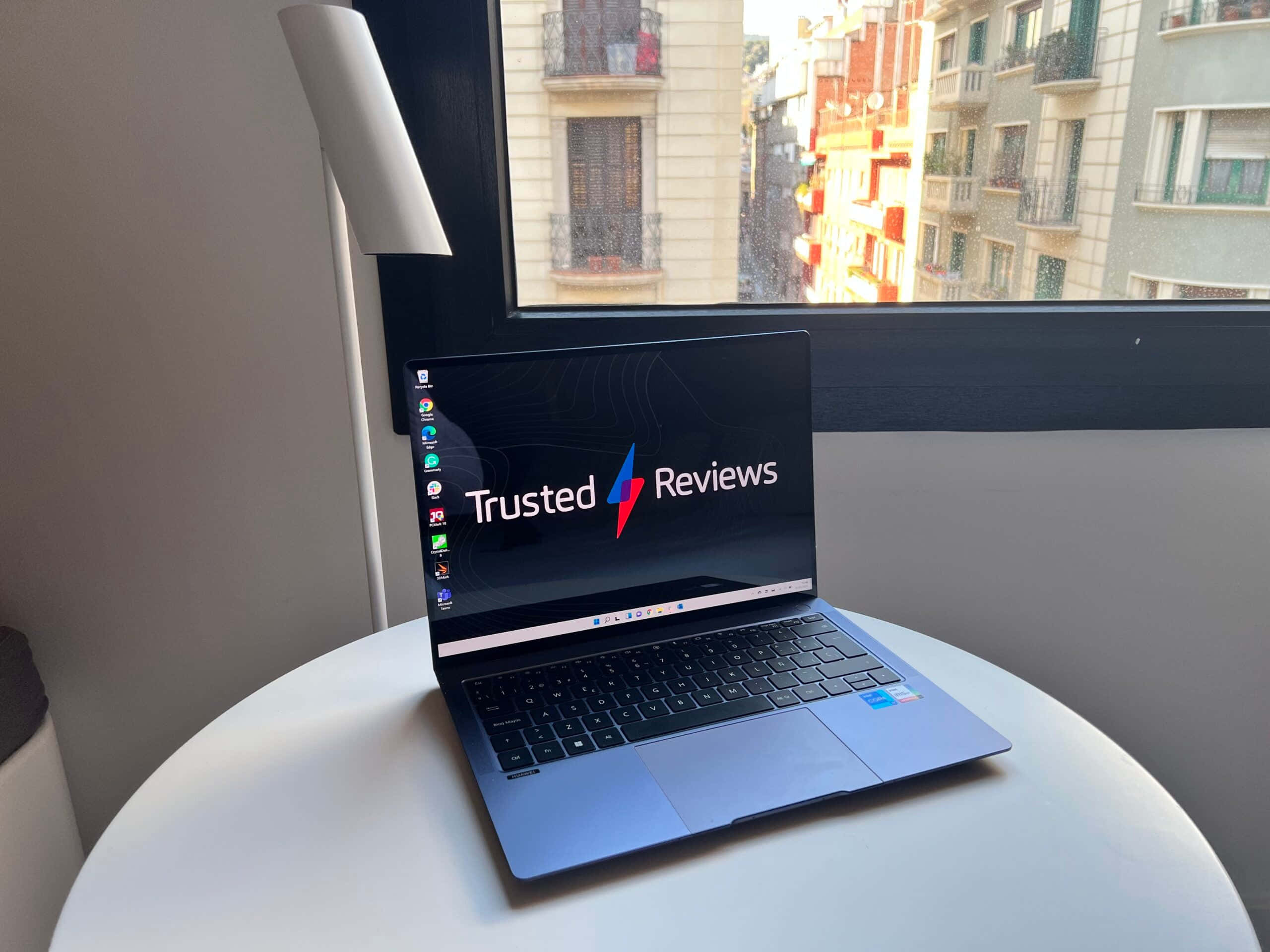 在家辦公 壁紙 — 一部時尚的藍色手提電腦顯示著 Trusted Reviews，放在靠近窗戶的白色圓桌上，窗外可俯瞰城市建築。