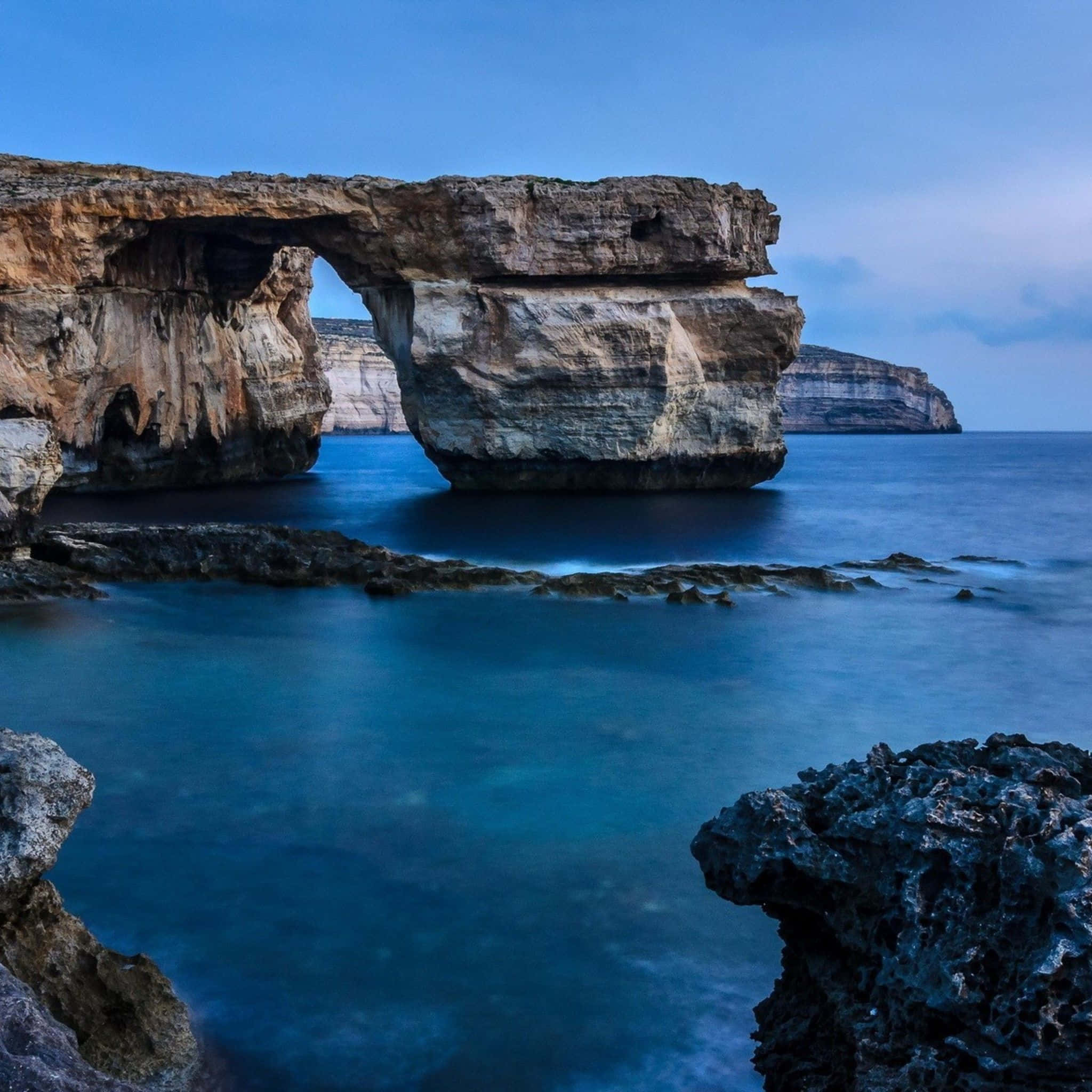 Malta 桌布 壁紙 — 在寧靜的暮色天空下，一座雄偉的自然岩石拱門矗立在平靜的碧綠海水之上。